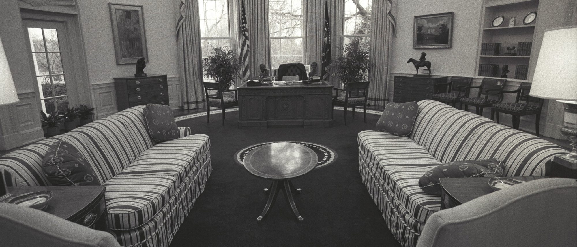 The Oval Office empty, photographed in black and white: the desk before the windows, two striped sofas facing each other and no one in the room