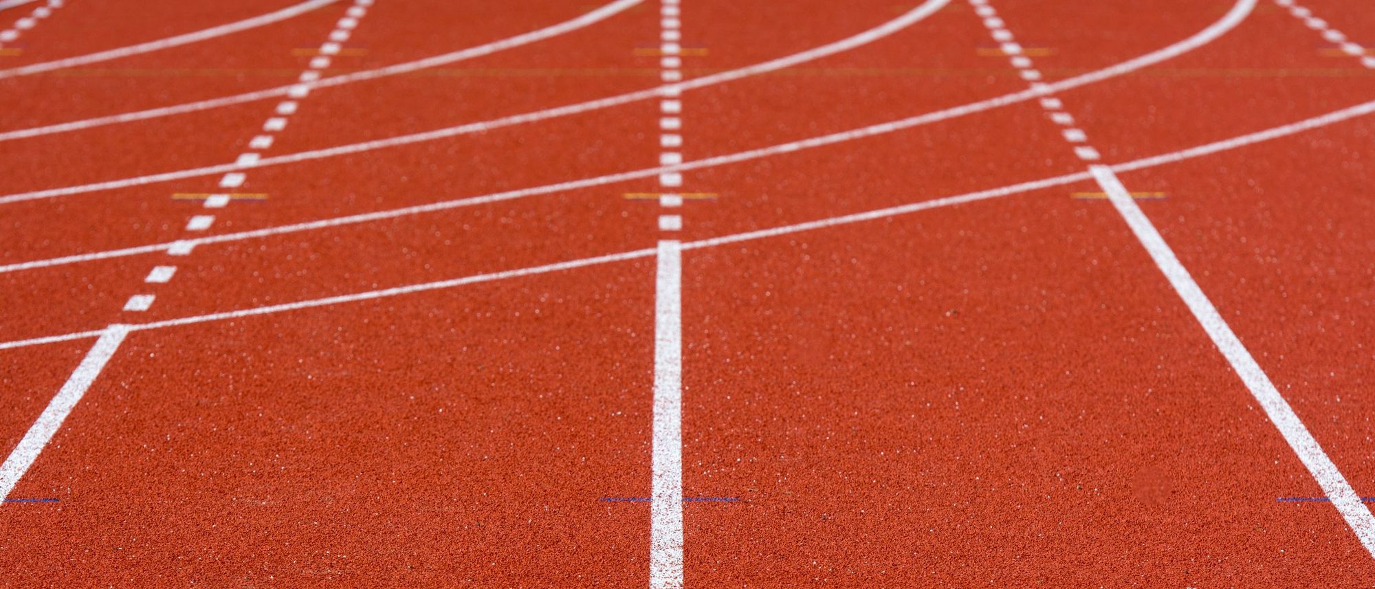 The red surface of an empty running track with white lane lines curving away.