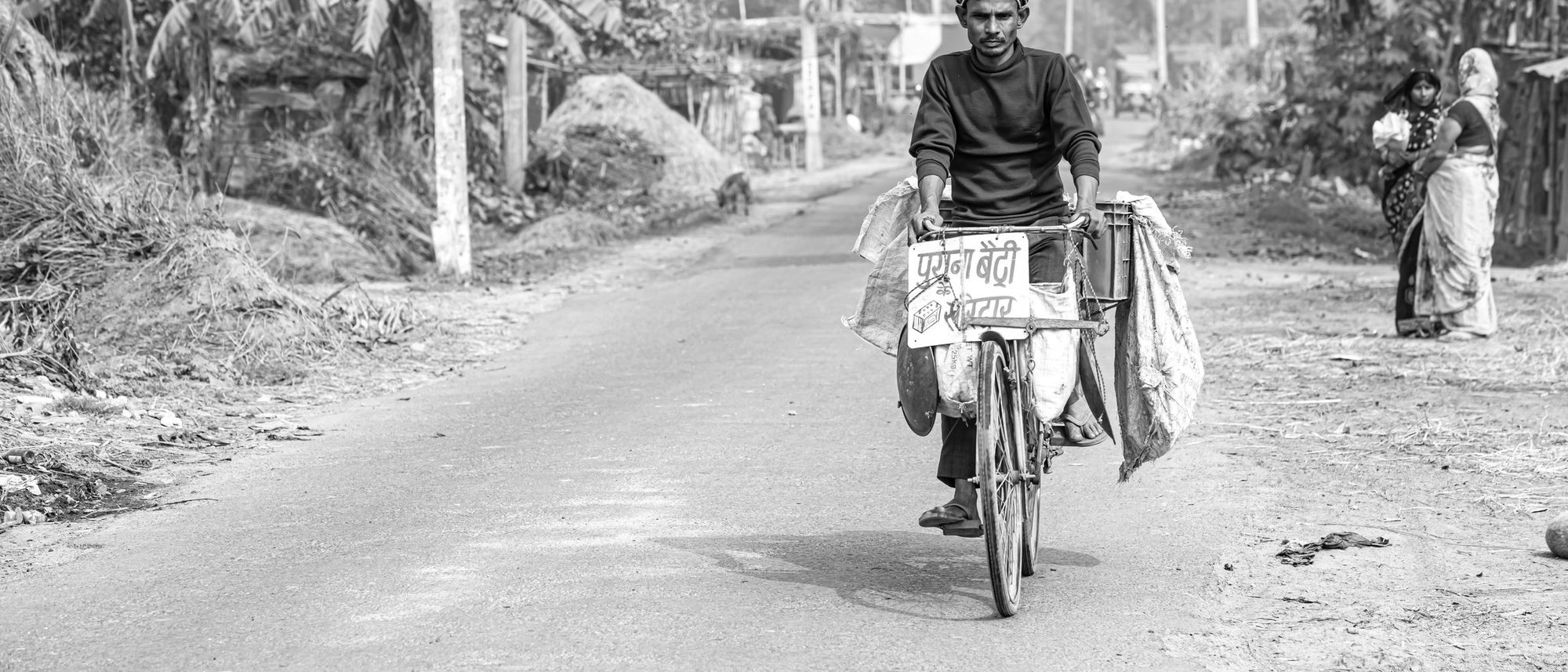 A man cycles along a village road in rural India with goods loaded on his bicycle.