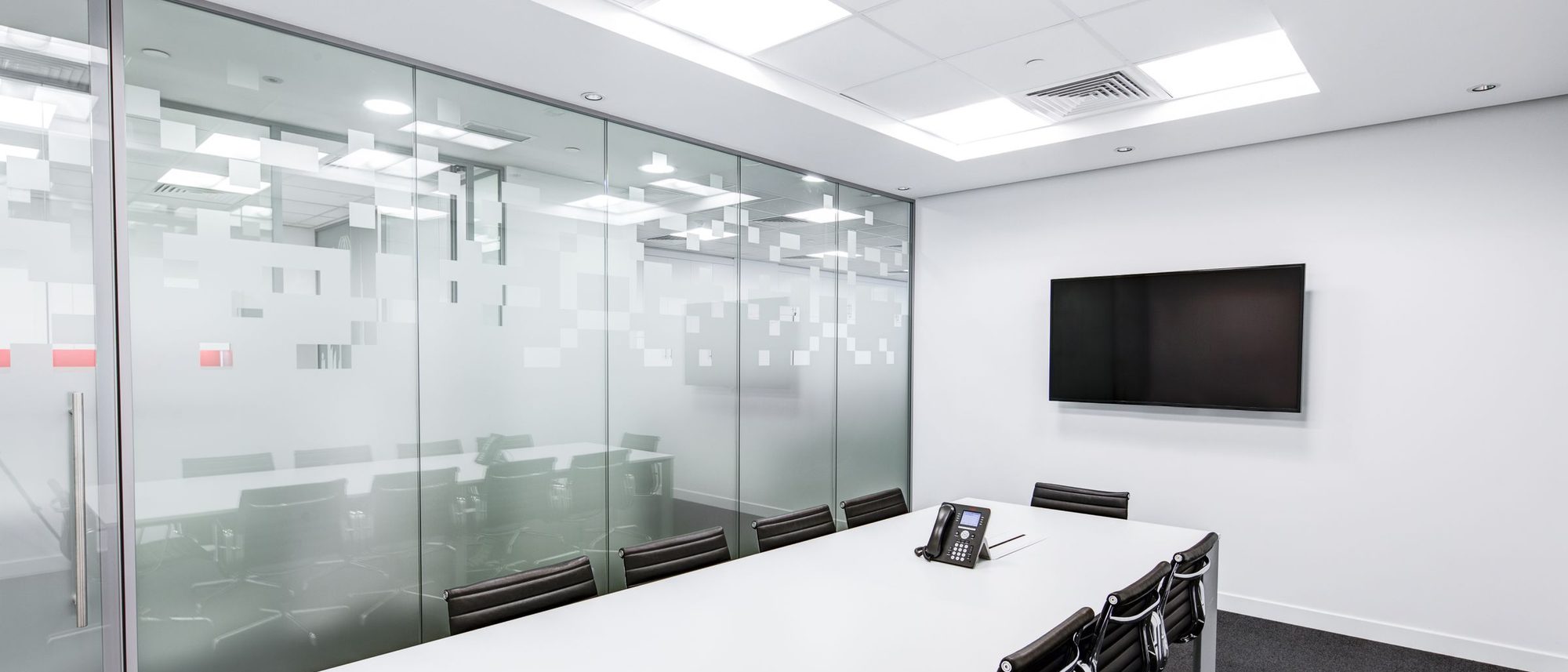 An empty glass-walled meeting room with a long white table and office chairs.