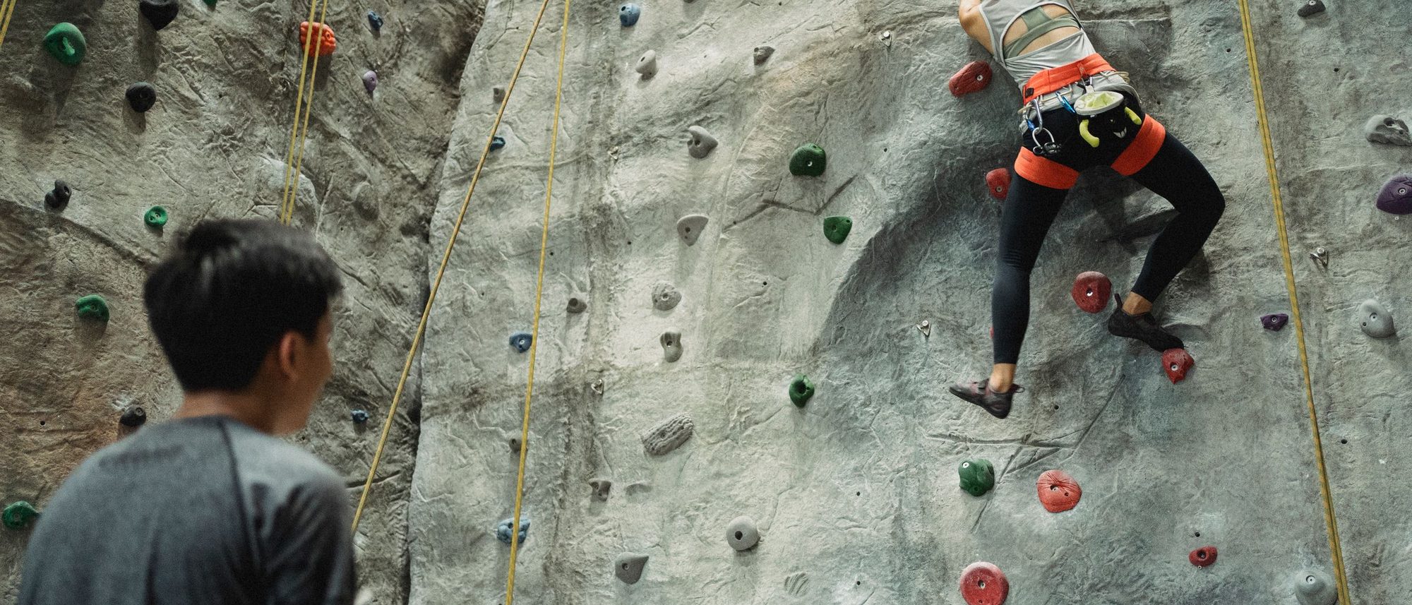 A climber on a grey indoor wall while a trainer watches from below, both seen from behind.