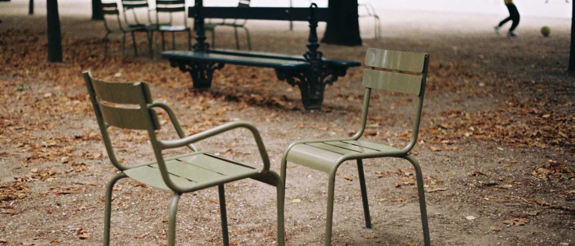 Two empty green metal chairs stand together in an autumn park with figures blurred behind.