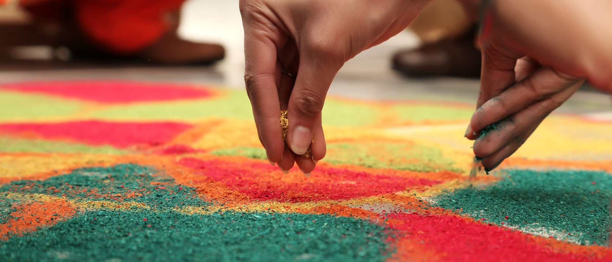 Hands sprinkle coloured powder to fill a rangoli pattern on the floor.