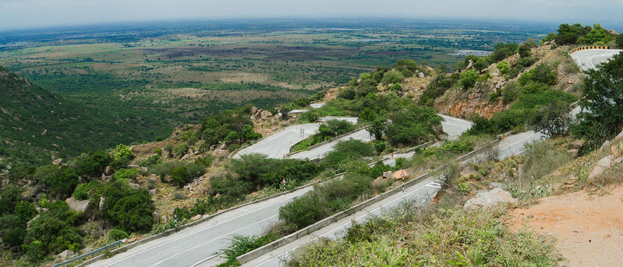 A road winds in tight curves through green hills in Karnataka under a cloudy sky.