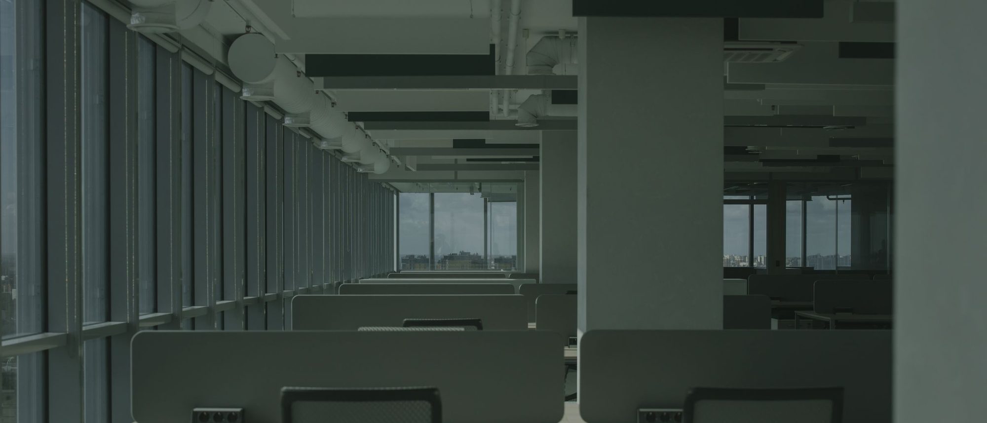 An empty office with rows of desks and large windows letting in daylight.