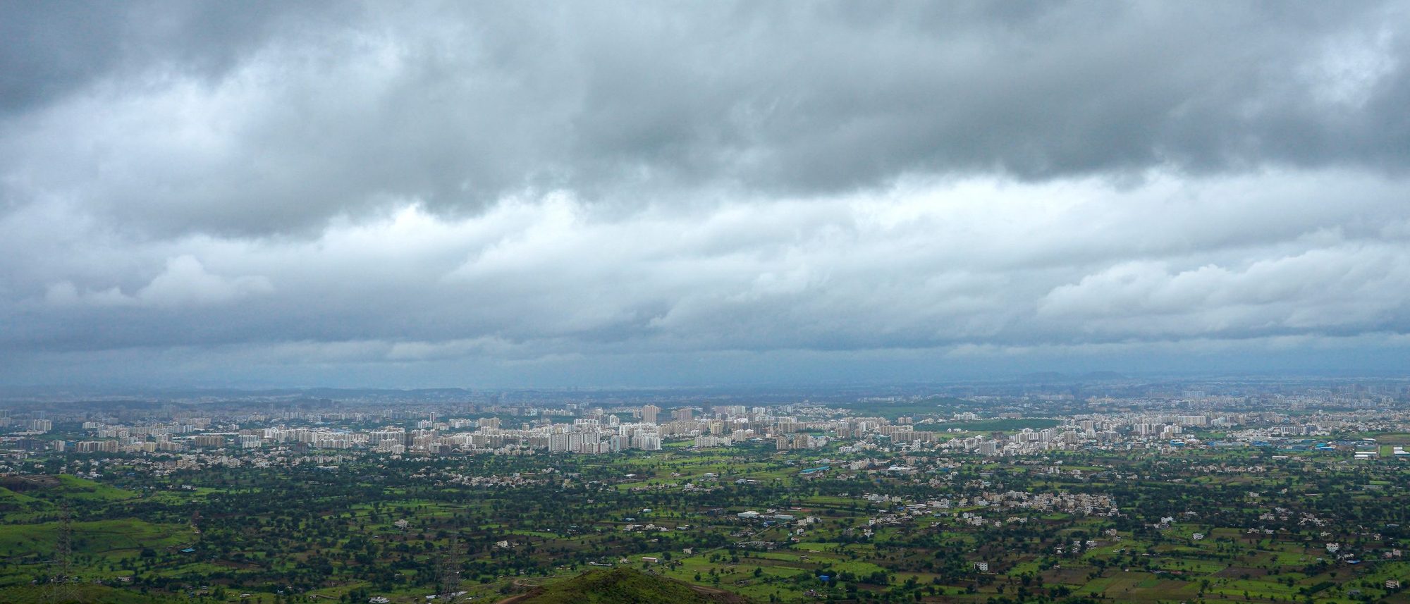 Aerial view of green fields and low hills at Holkarwadi in Maharashtra with a town beyond under a cloudy sky.