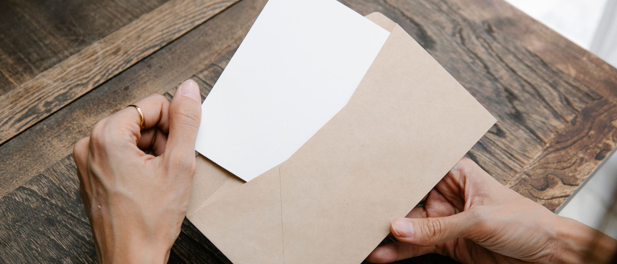 Seen from above two hands draw a blank sheet of paper out of a brown envelope on a wooden desk.