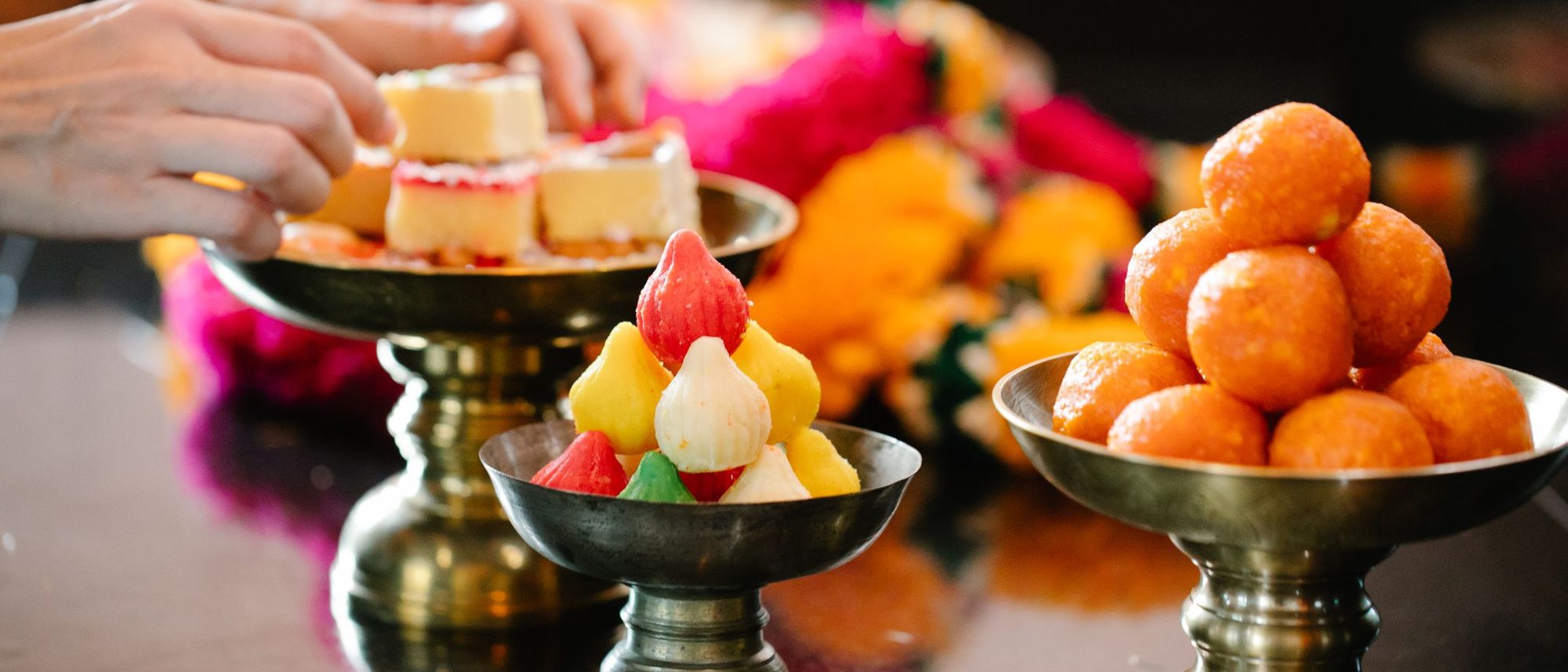 Colourful Indian sweets are piled on brass platters on a table.