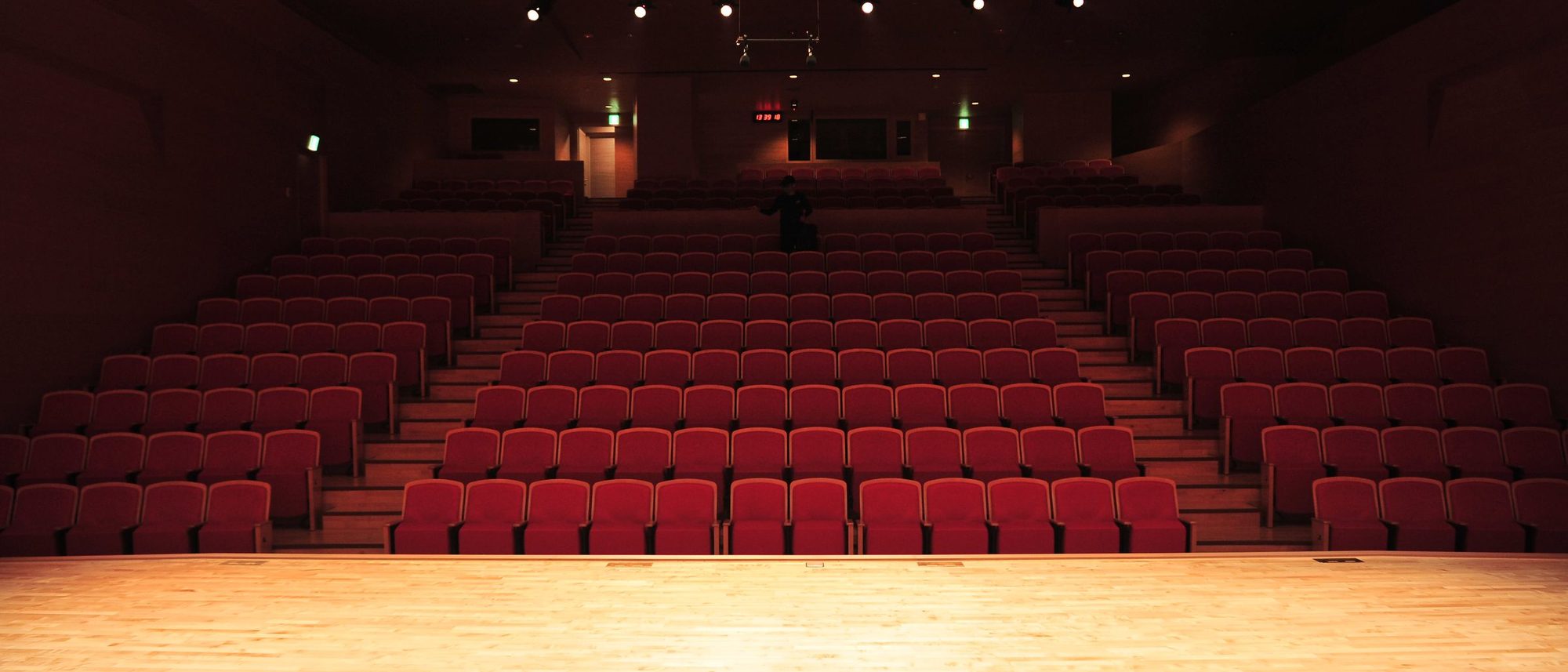 An empty theatre with rows of red seats faces a brightly lit stage.