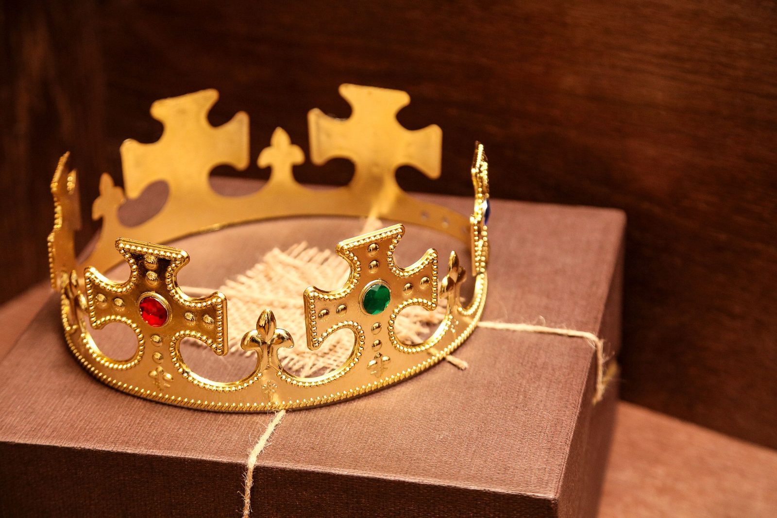 A gold costume crown set with red and green glass stones resting on a brown gift box tied with string