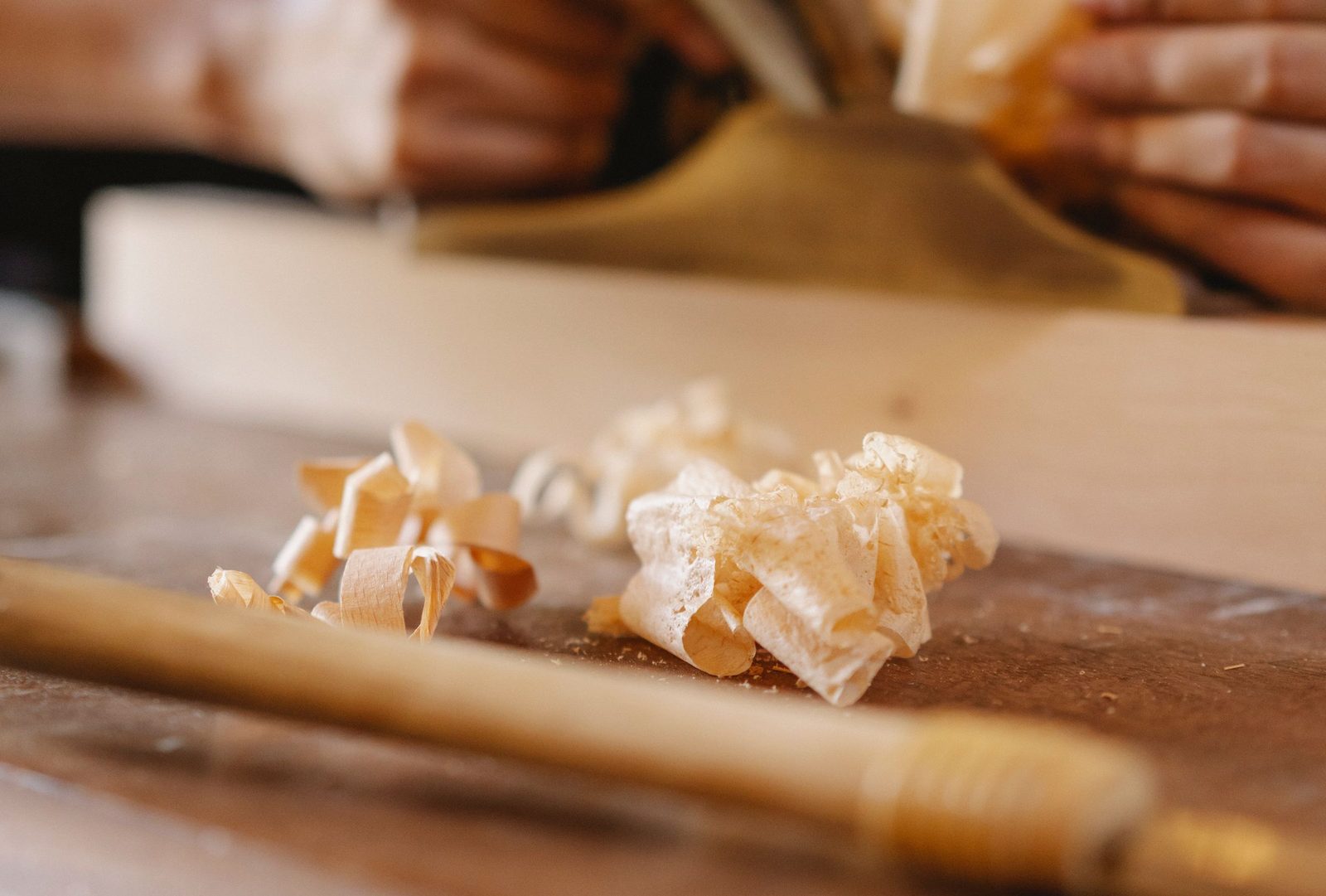 Thin curls of hardwood shavings on a workbench with a woodworker out of focus behind.