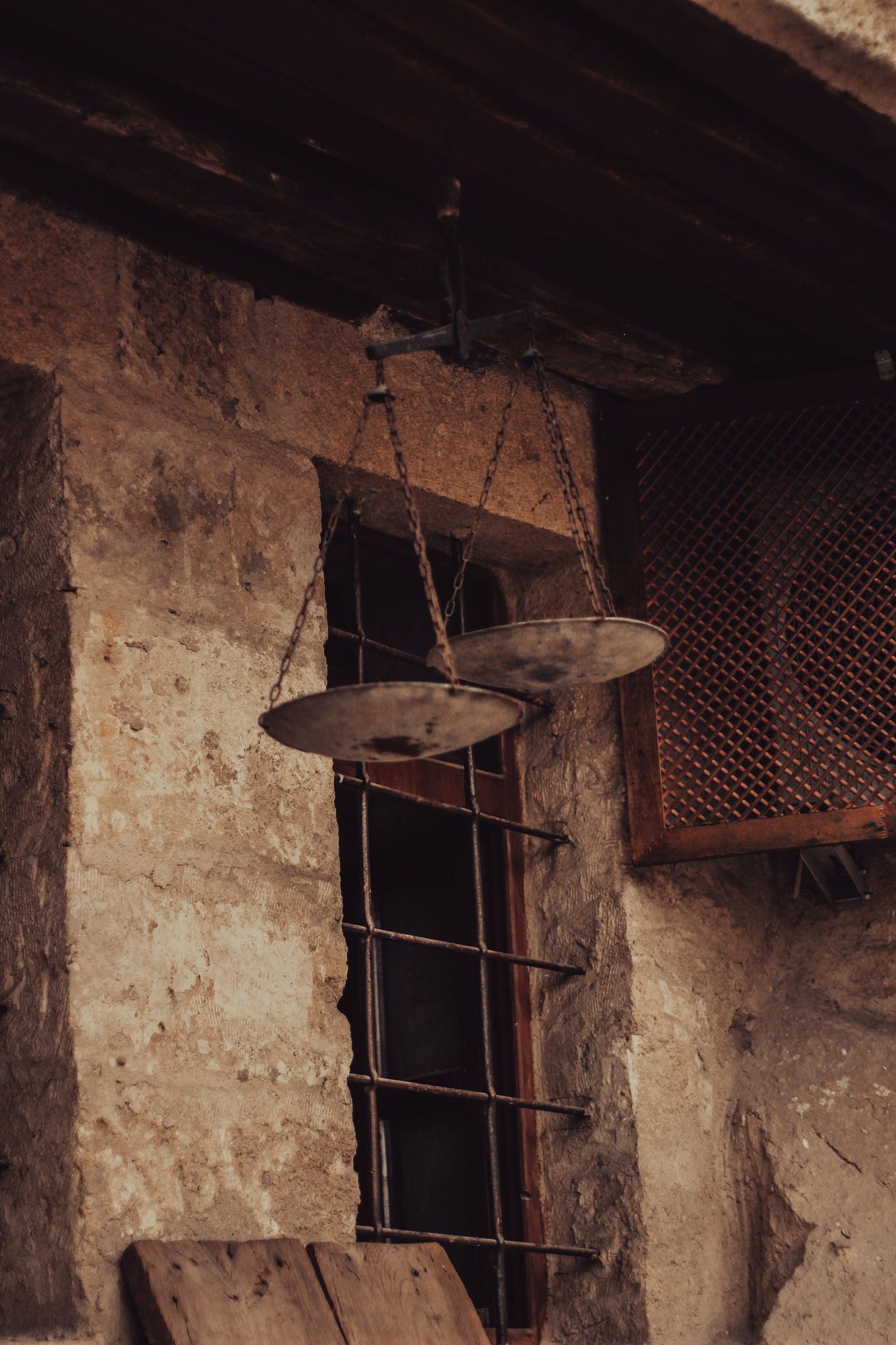 A weathered pan balance hangs on the wall of an old house beside a window.