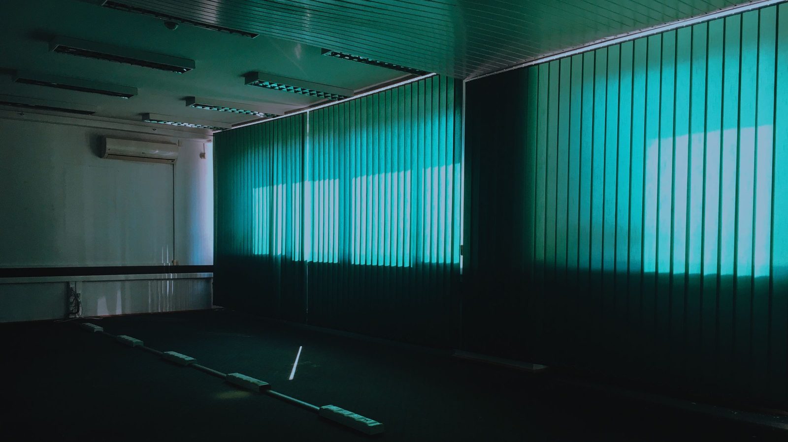 An empty office room in shadow with tall green blinds lit from behind by daylight