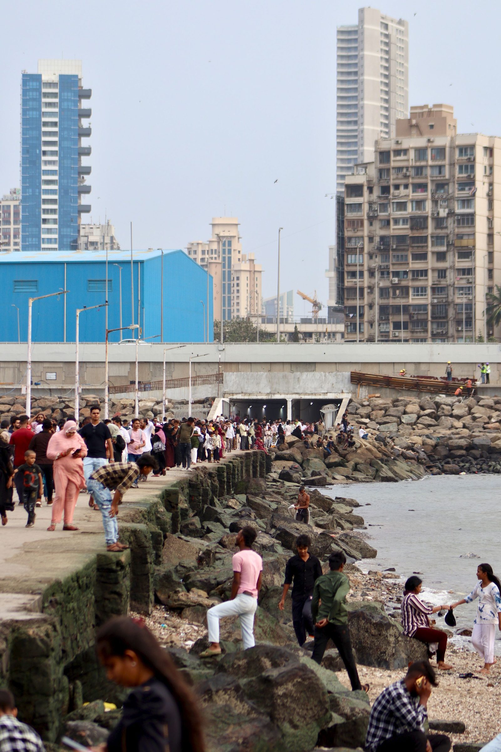 People walk along a crowded seafront promenade in Mumbai with tall buildings behind.