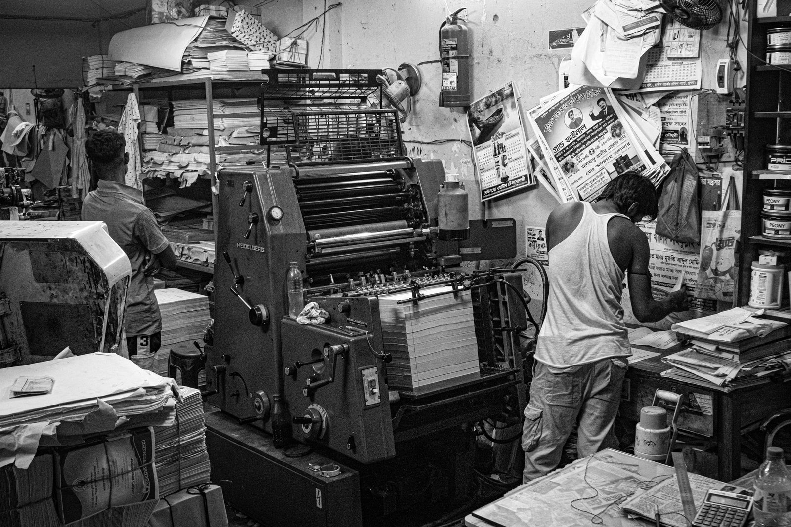 Workers operate a printing press in a busy print shop, in black and white.
