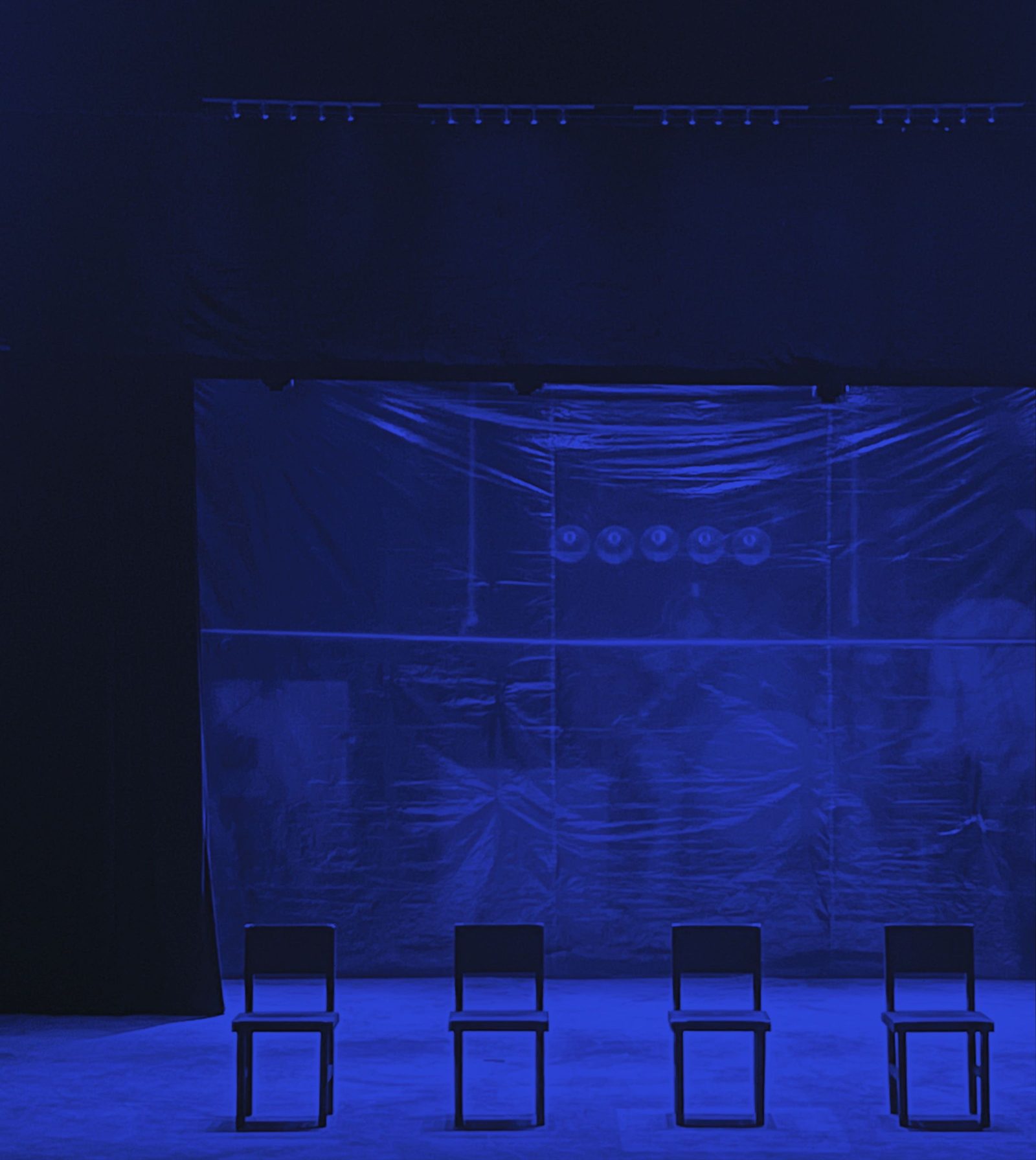 Four empty chairs on a dark stage under low blue light.