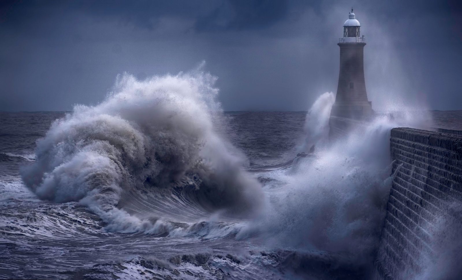 A heavy wave breaking against a lighthouse and sea wall on a stormy day.