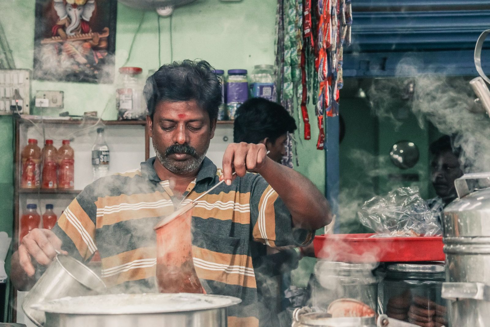 A man prepares masala chai at an Indian tea stall surrounded by steam and kitchen utensils.