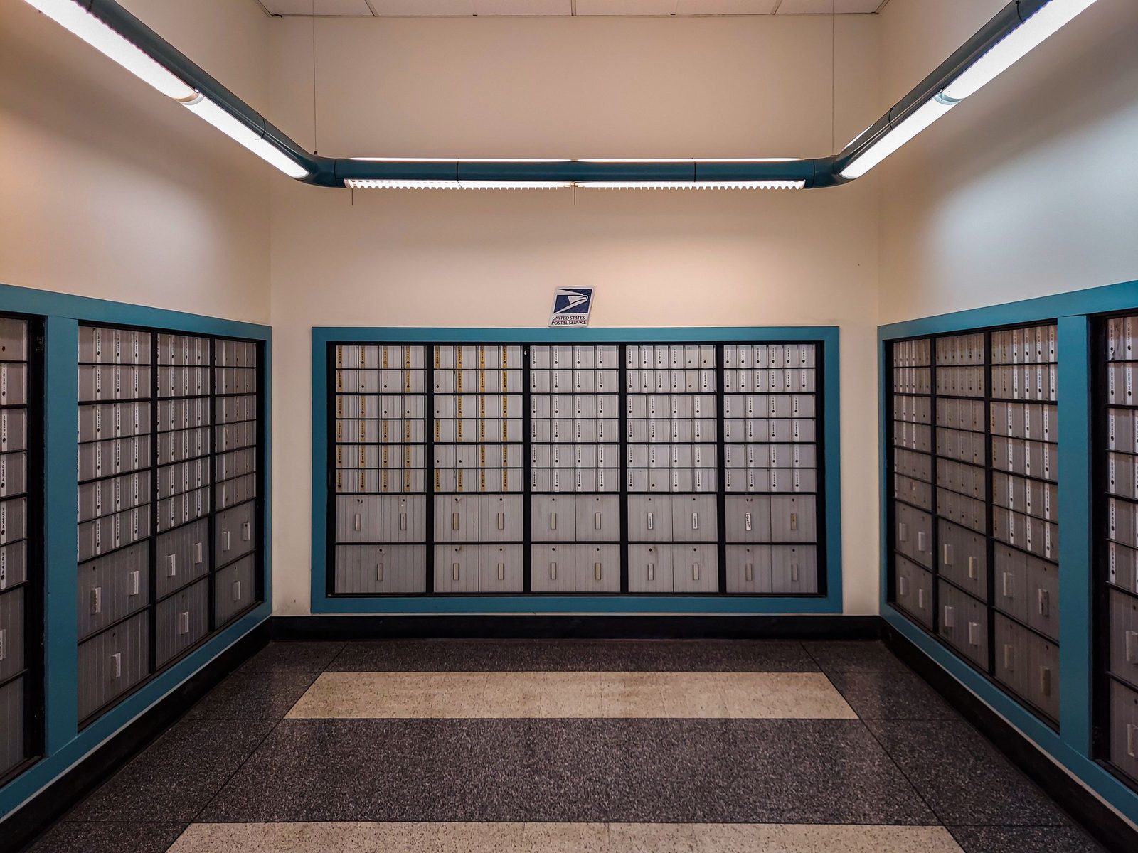 Rows of locked metal postal boxes line the wall of a post office under overhead lights.