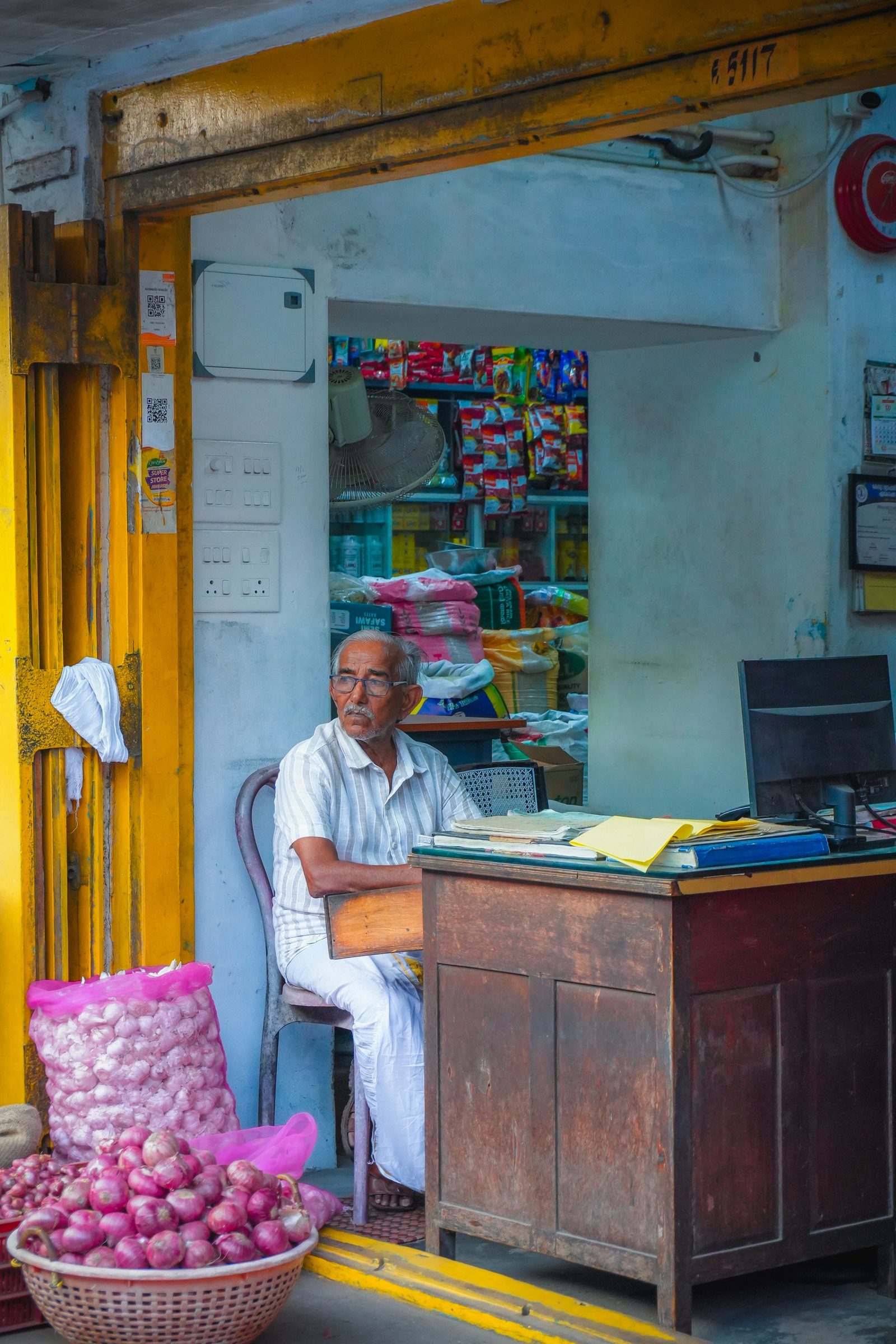 An elderly shopkeeper sits at a desk in a small grocery shop stocked with onions and packaged goods.