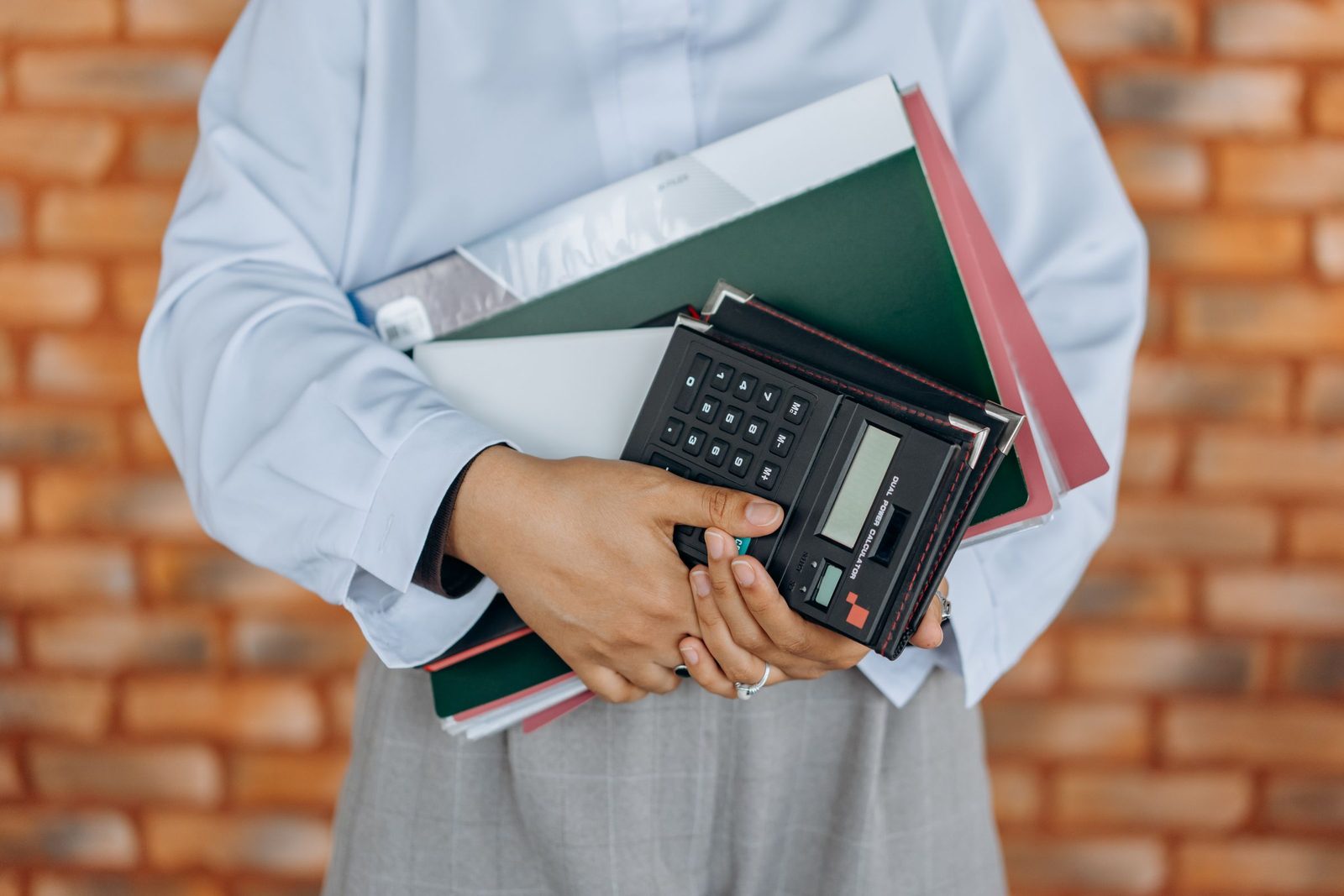 Hands holding a calculator and a stack of folders against a brick wall.
