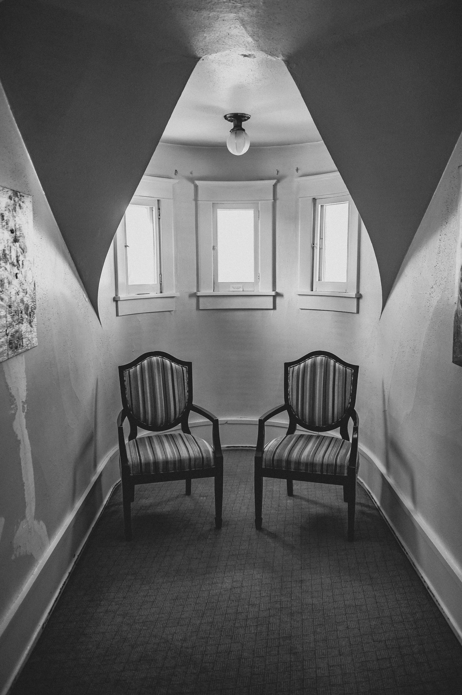 Two armchairs in a narrow room lit by tall windows, in black and white.