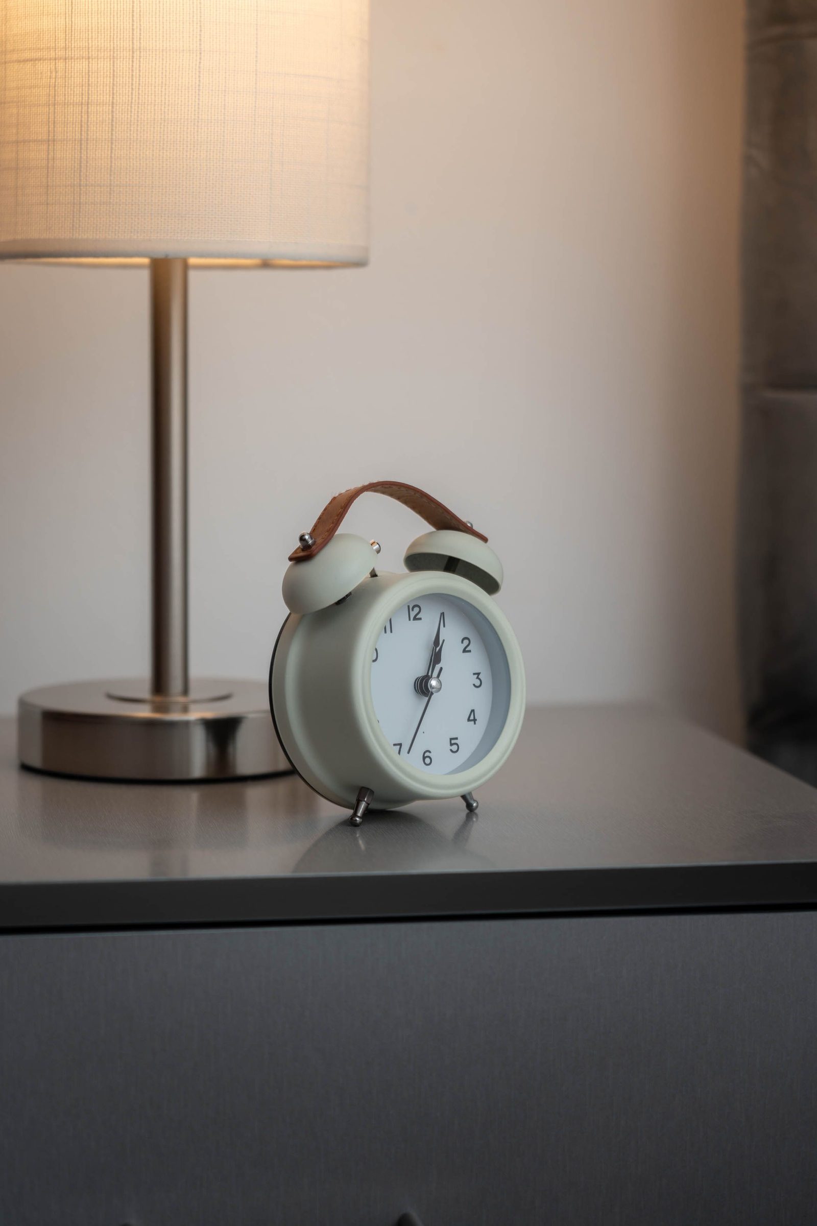 An analogue alarm clock sits beside a lit lamp on a bedside table.
