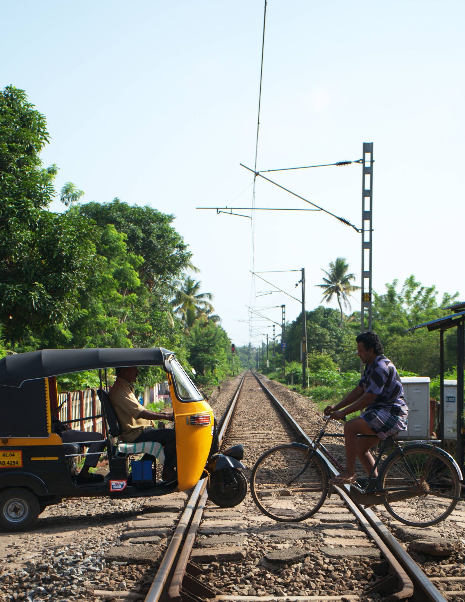 An auto rickshaw and a cyclist waiting at a rural railway crossing in Kerala under a clear sky.
