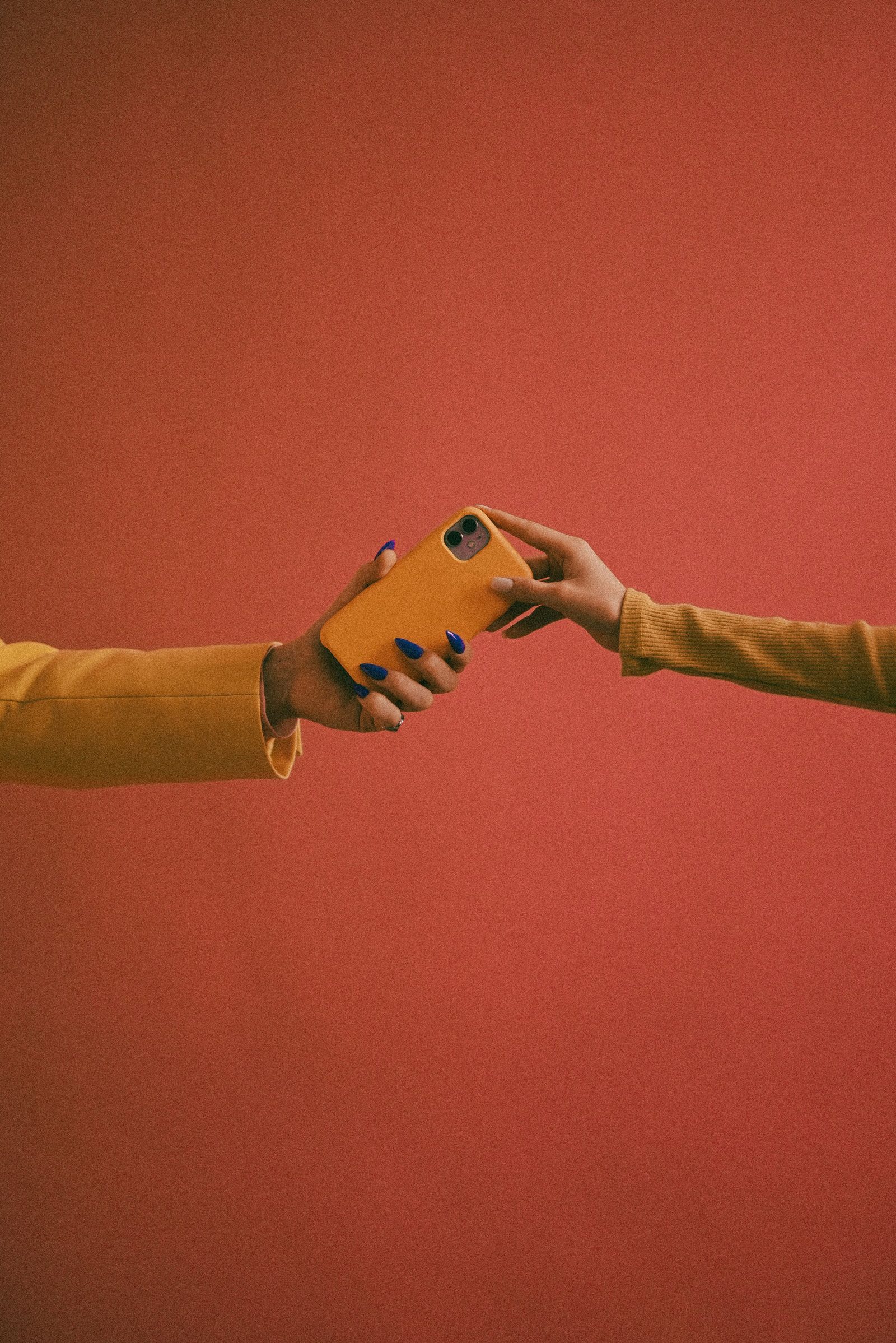 Two hands pass a smartphone between them against a bright orange wall.