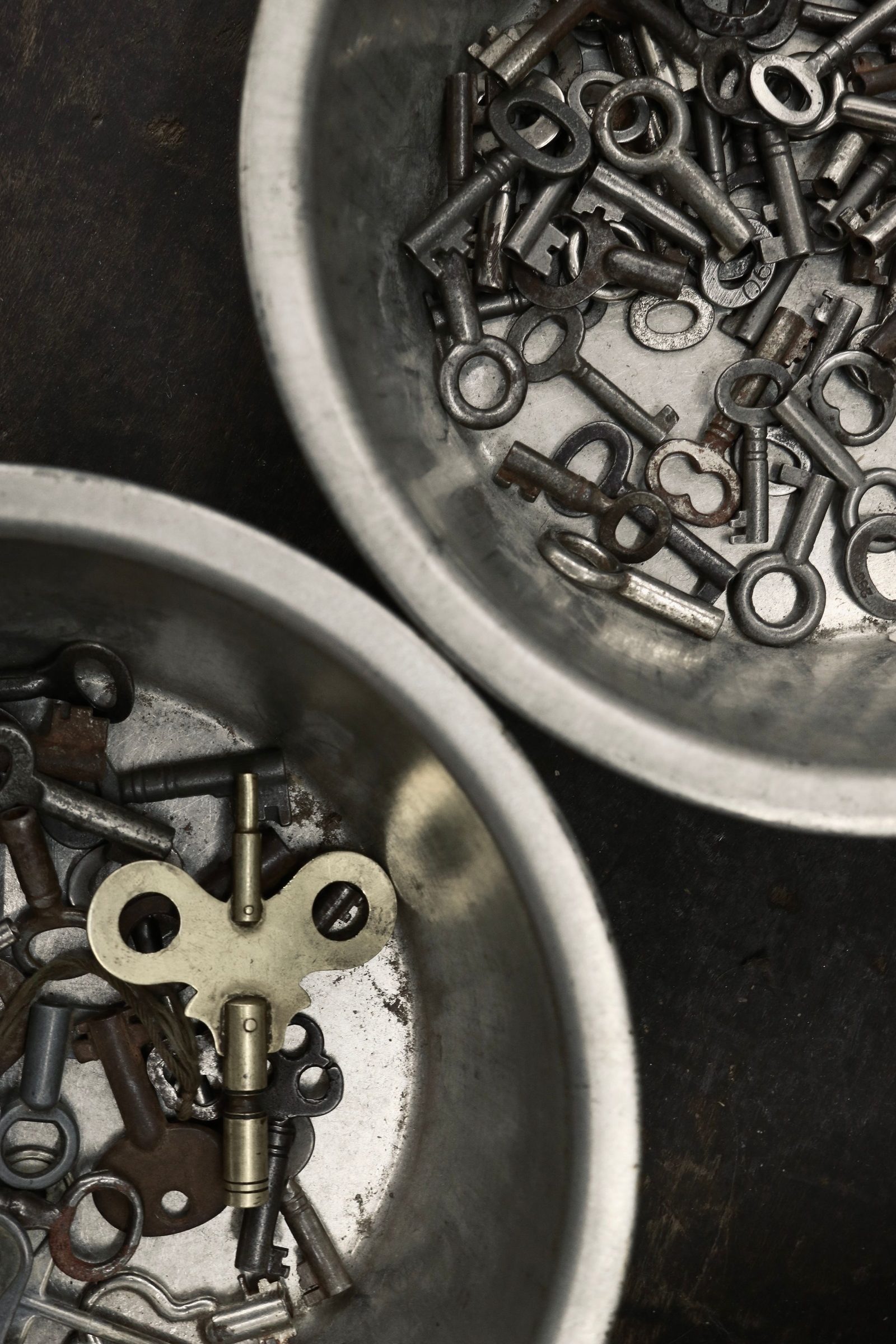 Two metal bowls full of assorted old keys on a dark surface, seen from above.