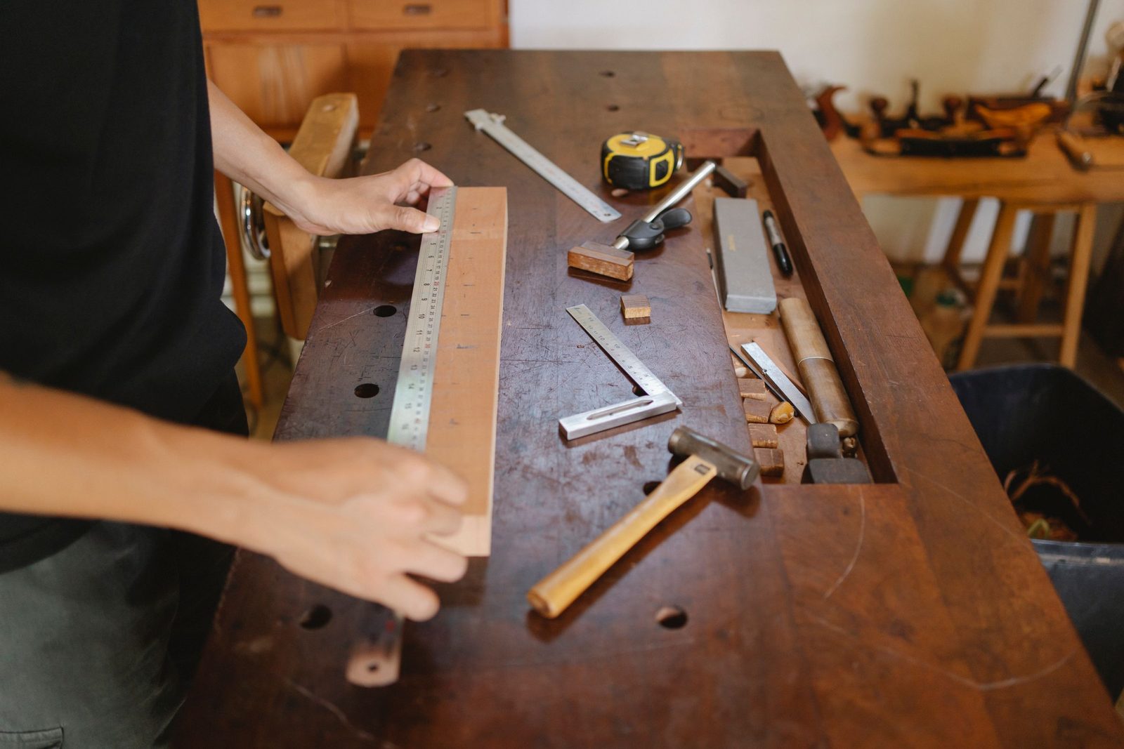 Hands measuring a timber plank with a ruler on a carpenter’s bench beside a hammer and tools.