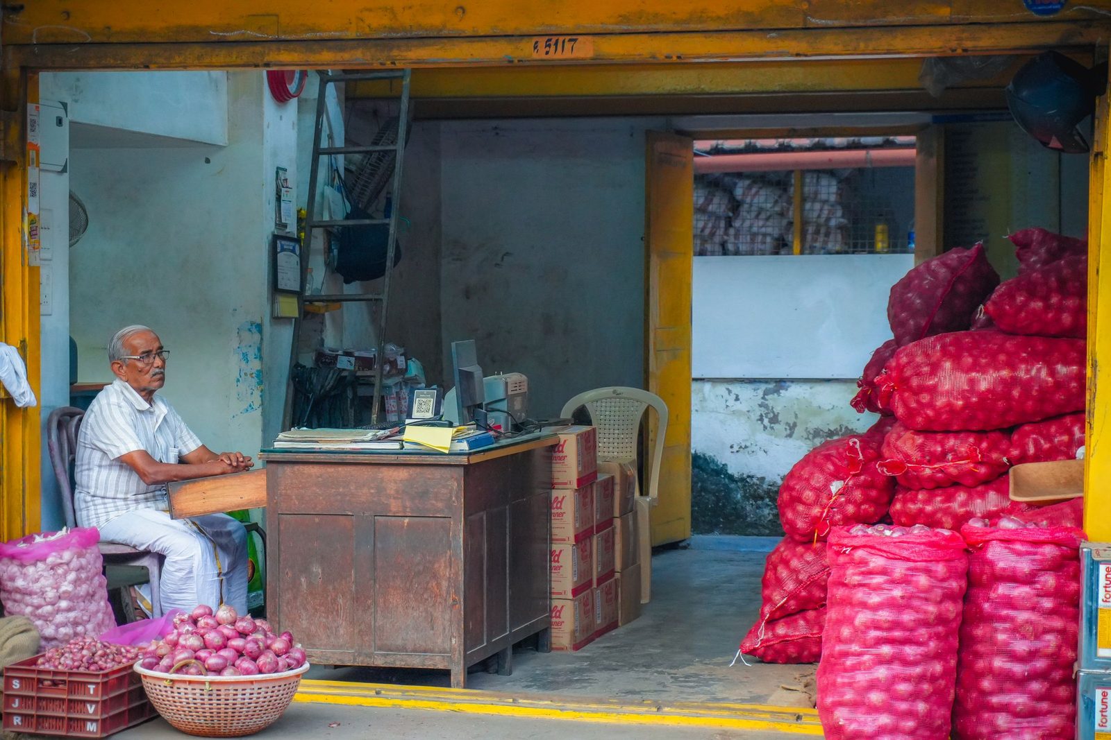 A shopkeeper sits in a small market shop among sacks of onions and stacked boxes.