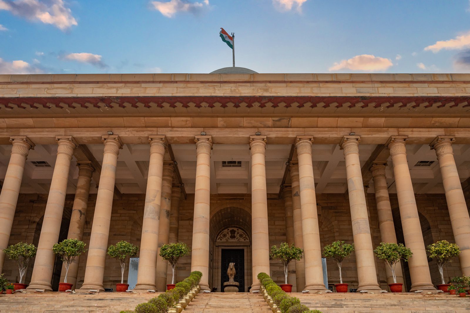 A colonnaded government building in New Delhi with the Indian flag against a bright sky.