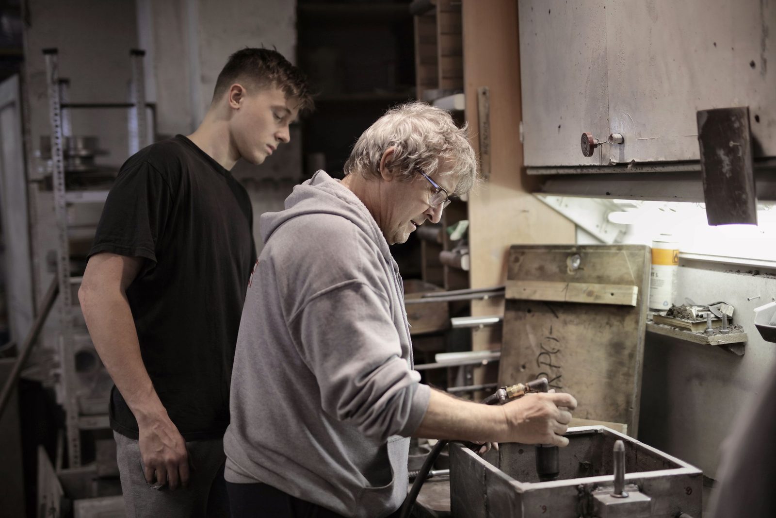 An experienced craftsman shows a young apprentice how to handle a part at a workbench.