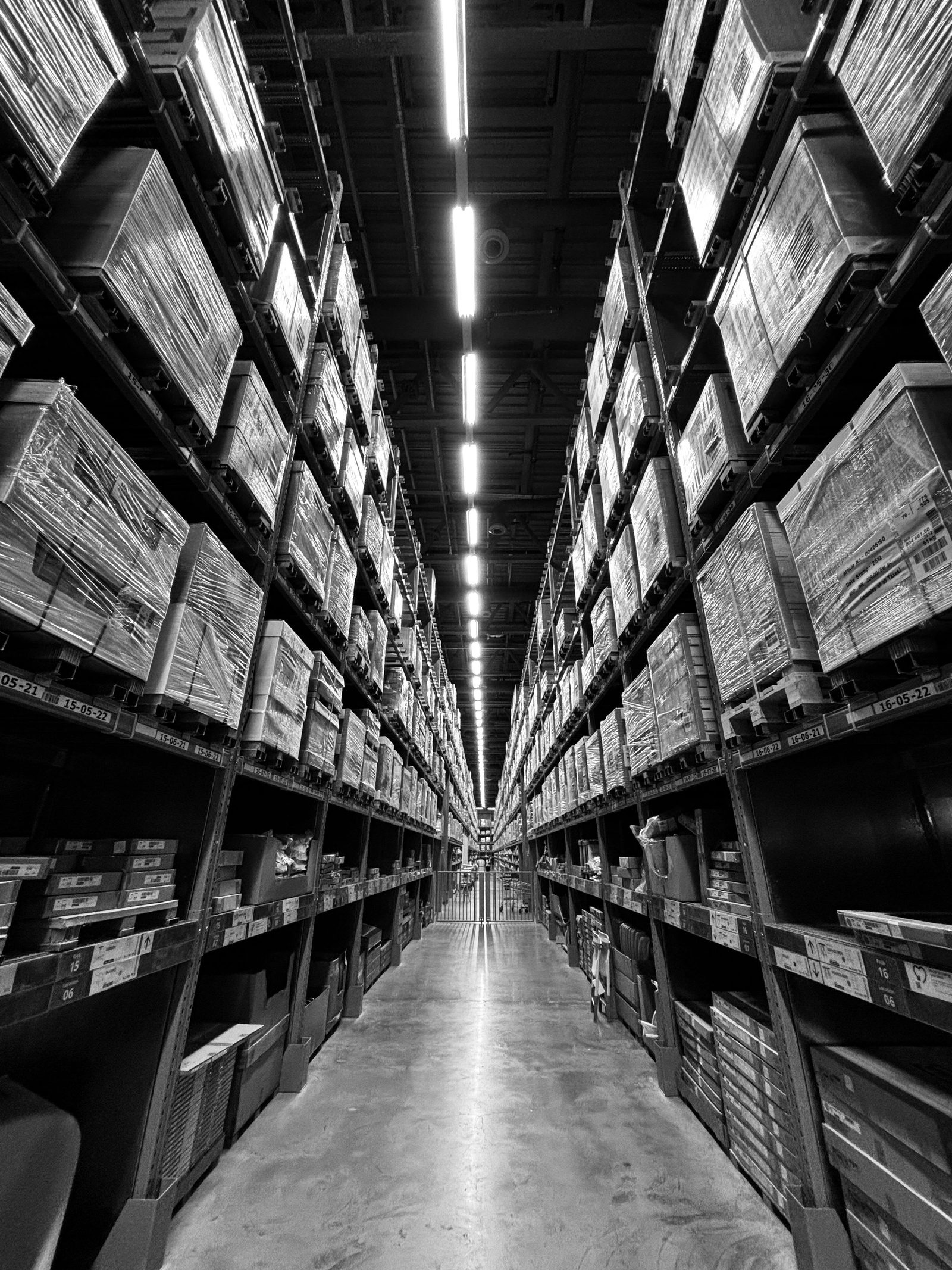 A black and white view down a warehouse aisle with boxes stacked high on both sides.