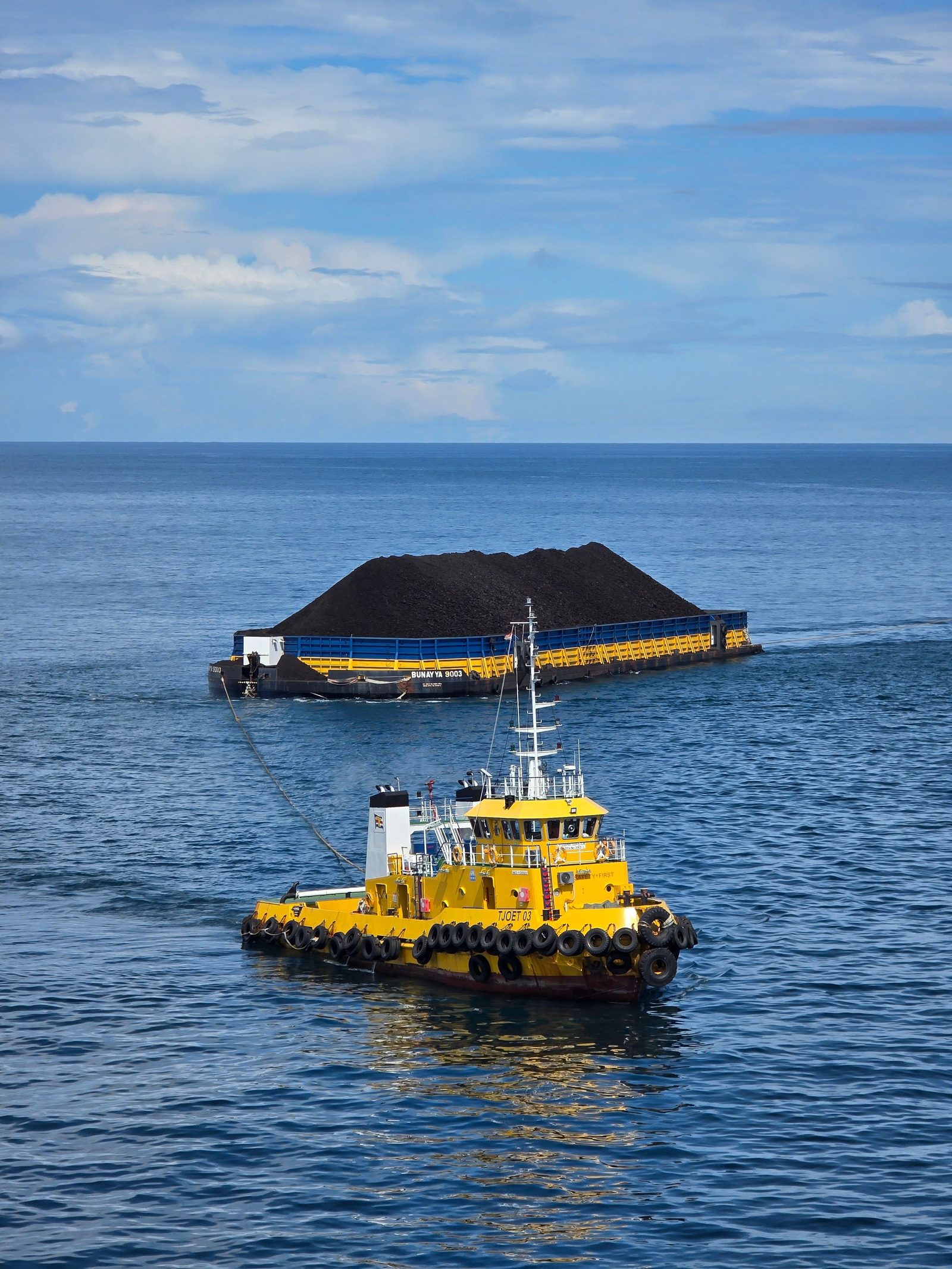 A yellow tugboat tows a loaded coal barge across a calm blue sea.