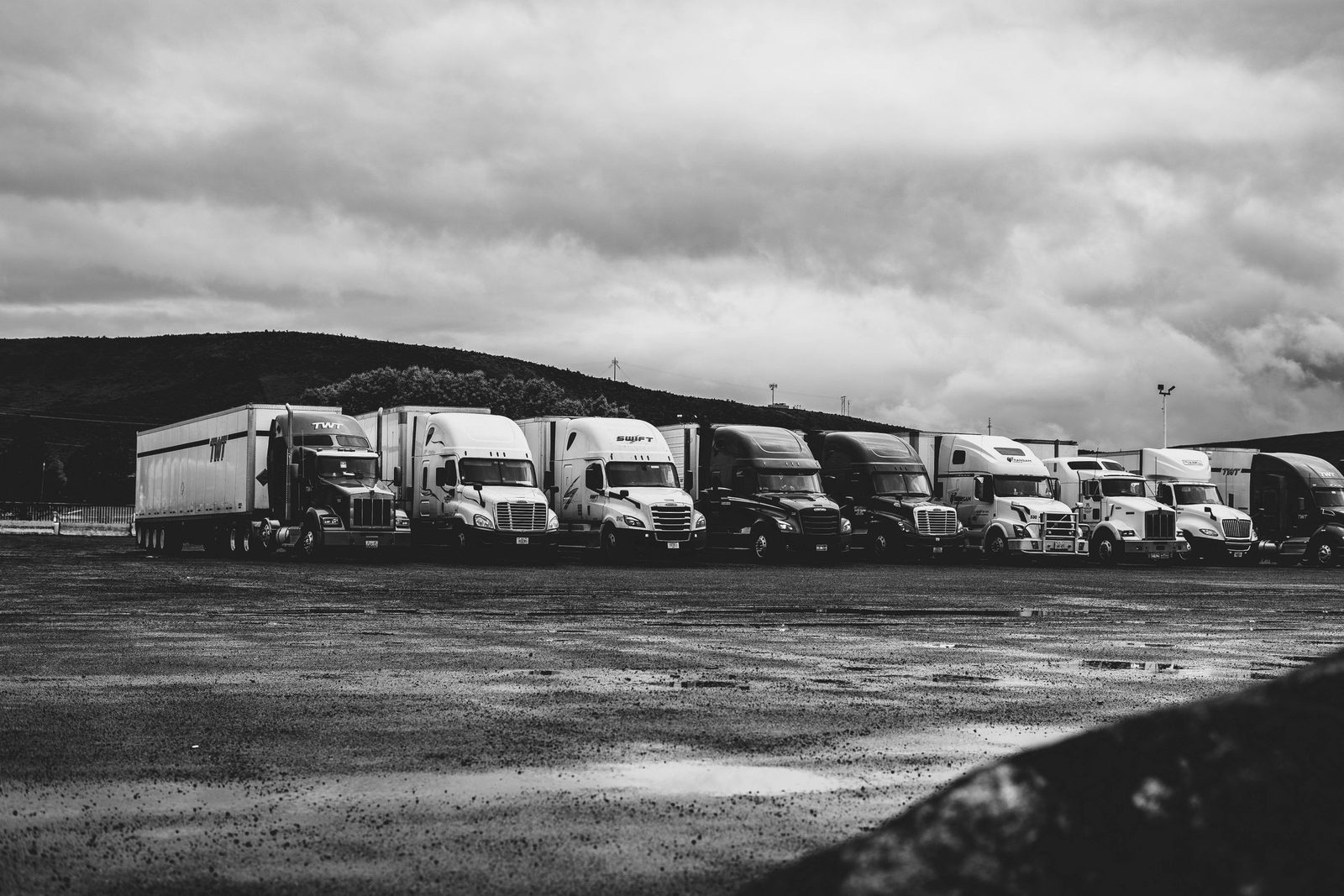 A row of parked lorries in a wet yard under heavy cloud, in black and white.