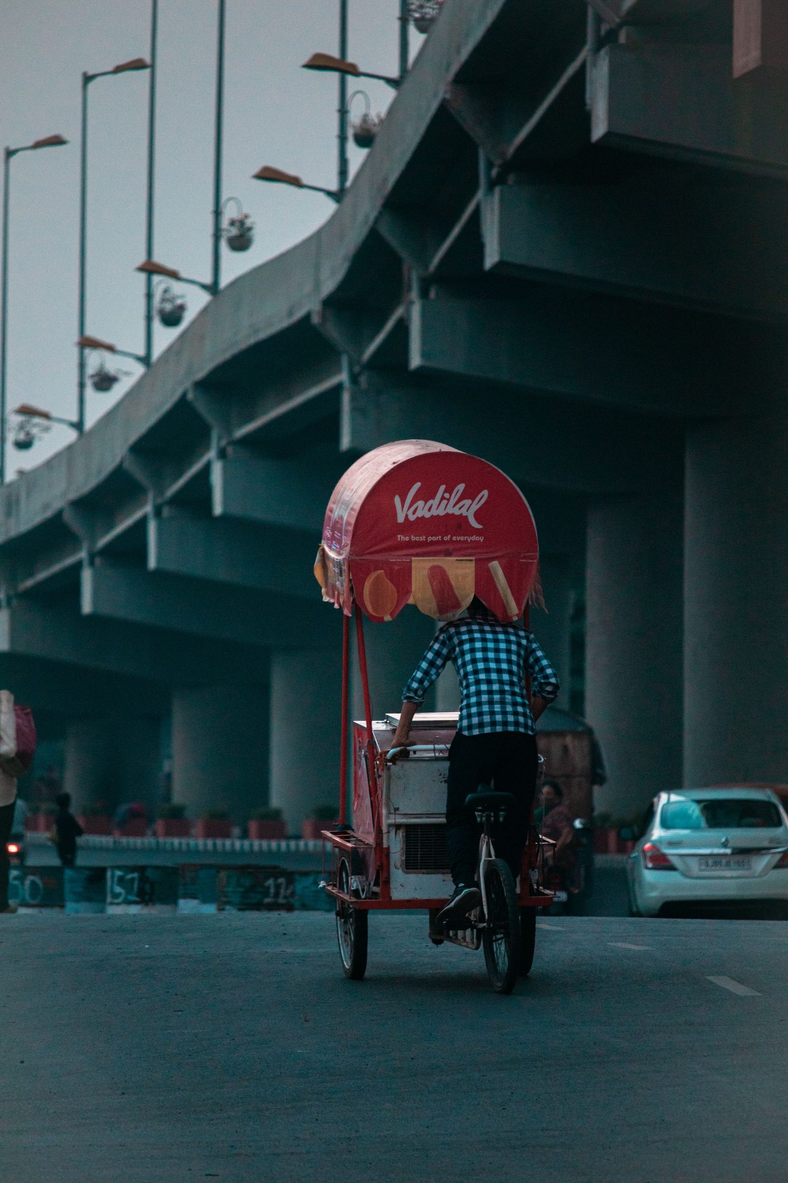 A vendor pedals a loaded cargo bicycle beneath a flyover on a street in Surat.