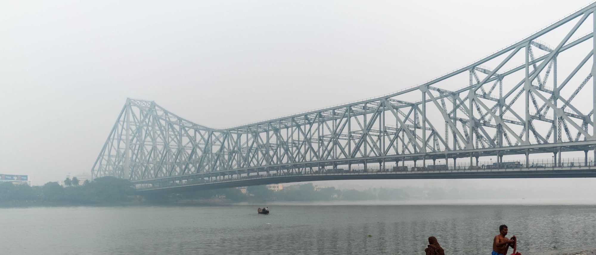 Howrah Bridge emerges from morning mist over the river with small figures on the bank.