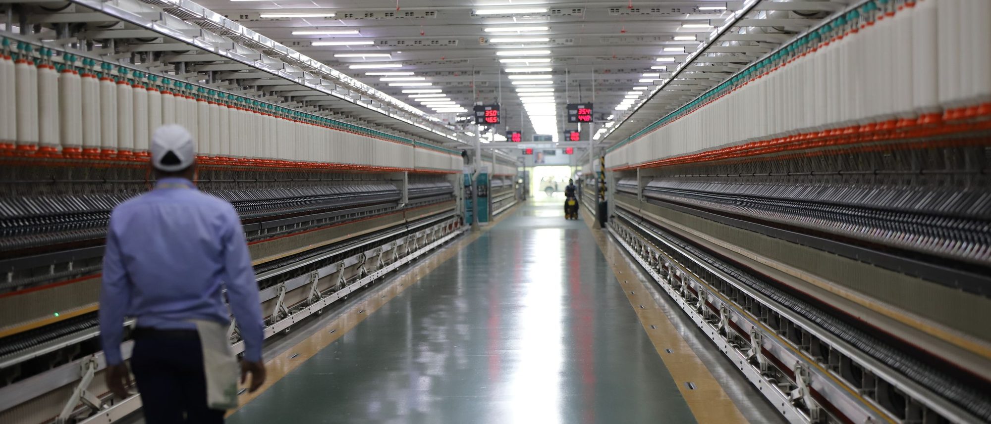 A long aisle between spinning machines in an Indian textile mill, with a single worker walking away down it.