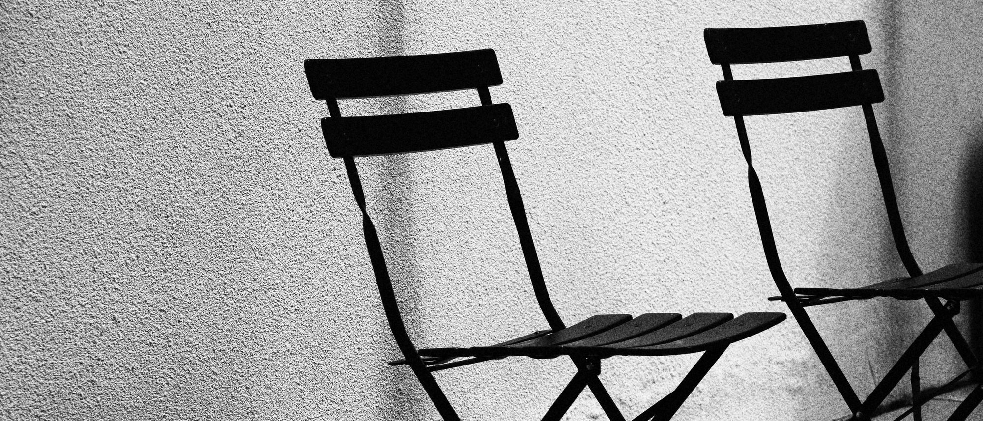 Two simple chairs side by side against a textured wall casting long shadows, in black and white.