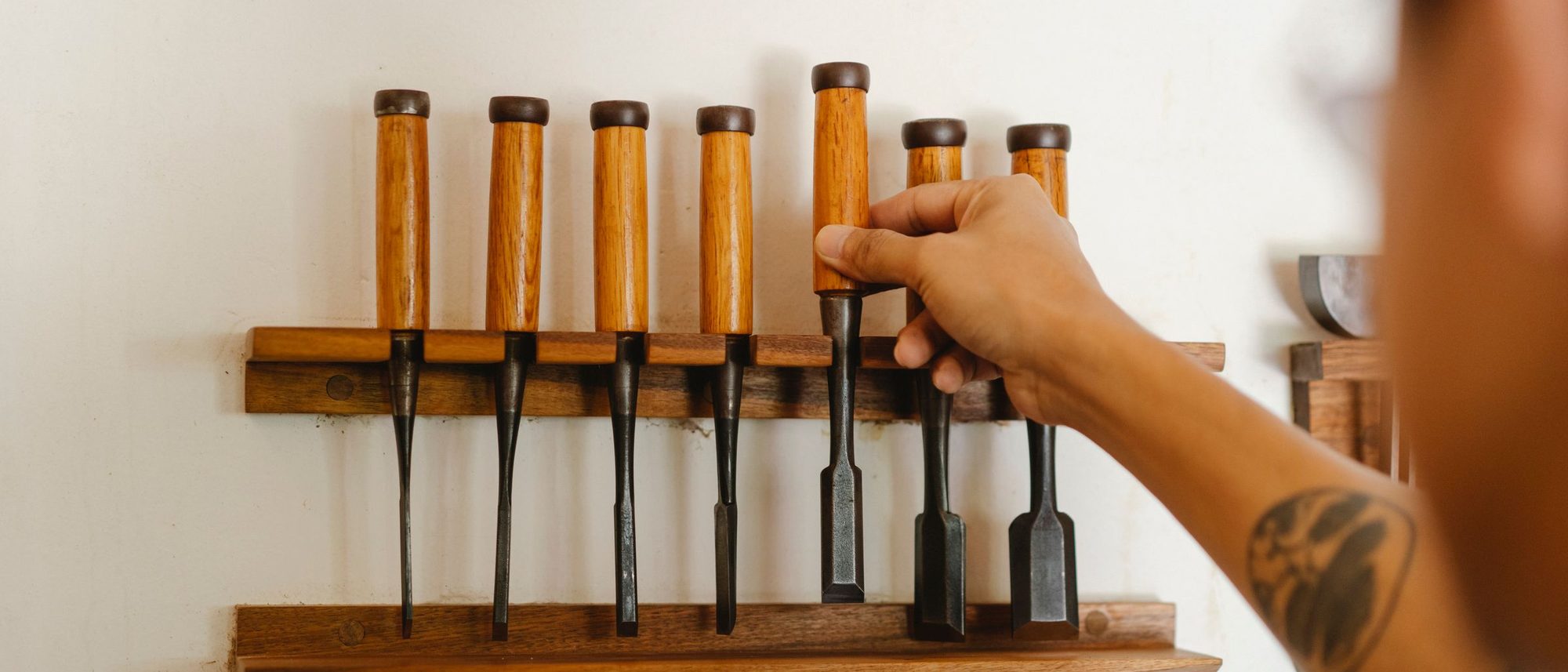Seen from behind a joiner reaches for one chisel from a neat row of tools on a workshop wall.