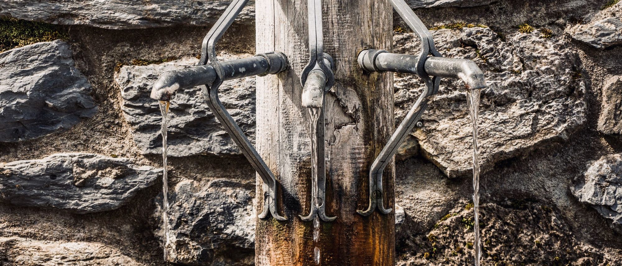 Metal taps pour water into a stone trough beside an old wall on a city street.
