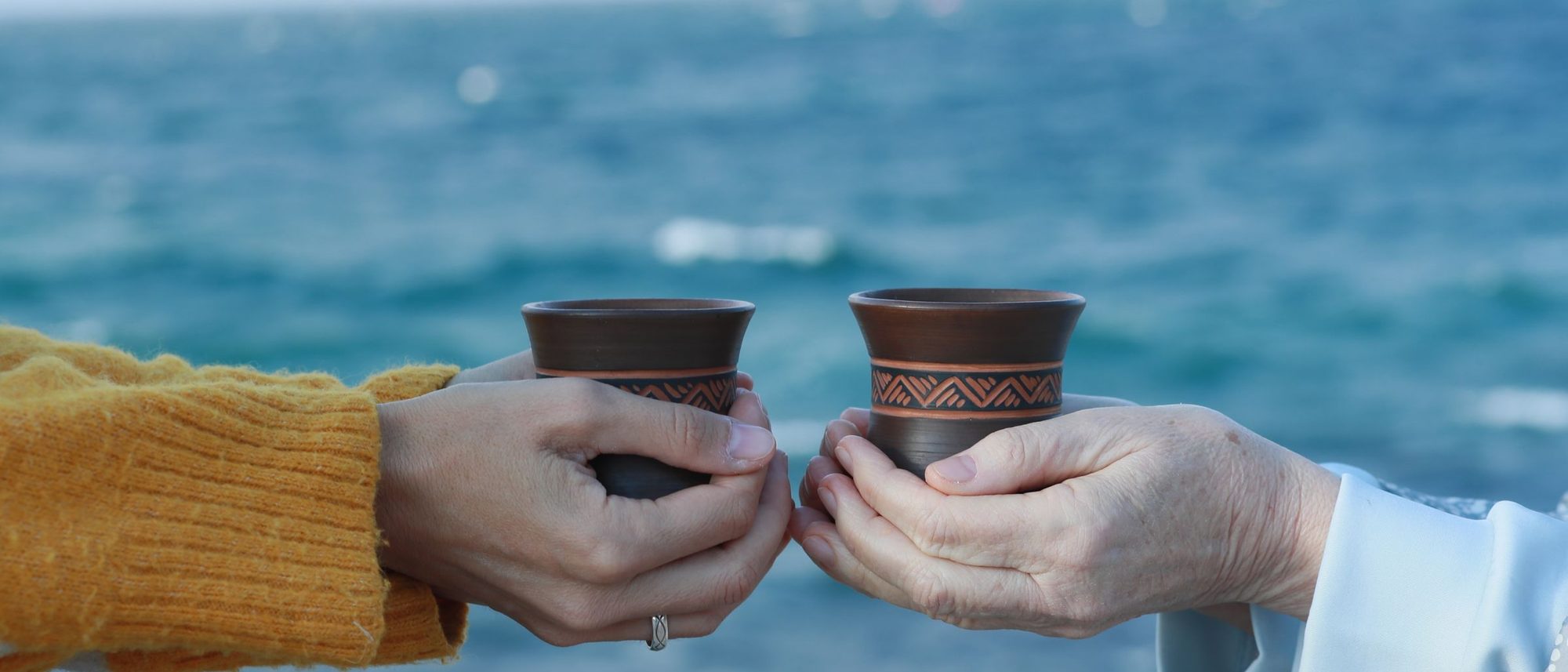 Two people hold clay cups of tea side by side by the sea.