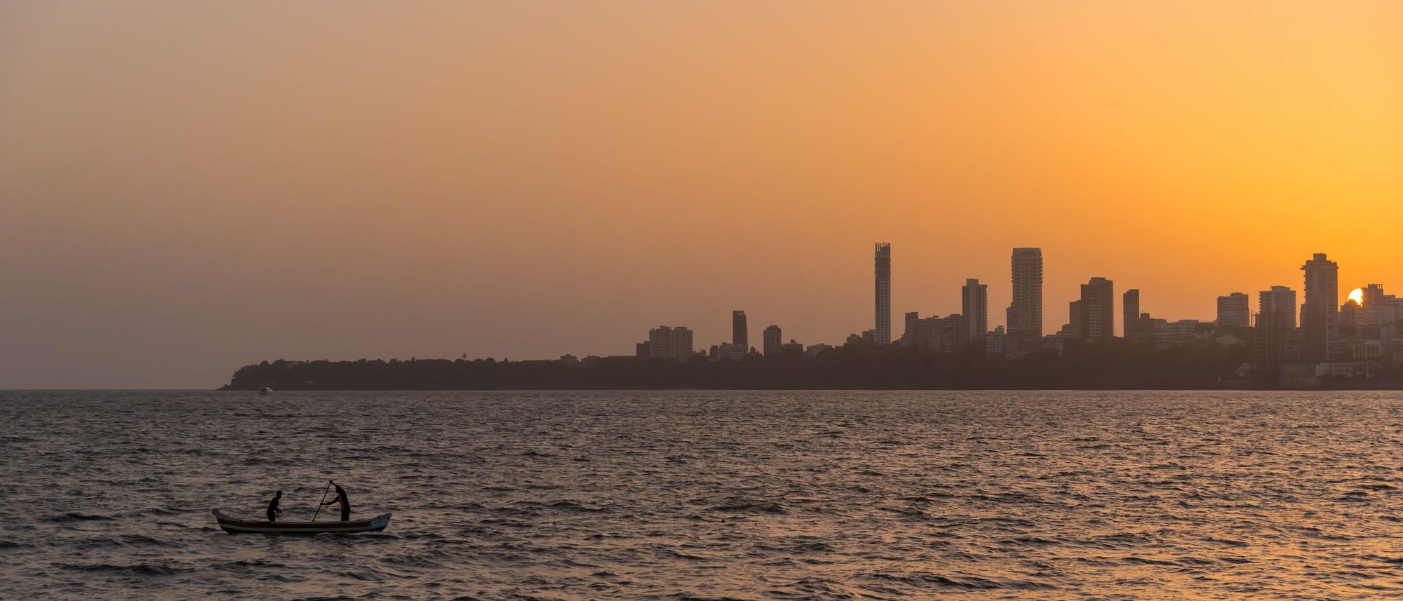 Two fishermen in silhouette on a small boat at sunset, with the Mumbai skyline behind them.