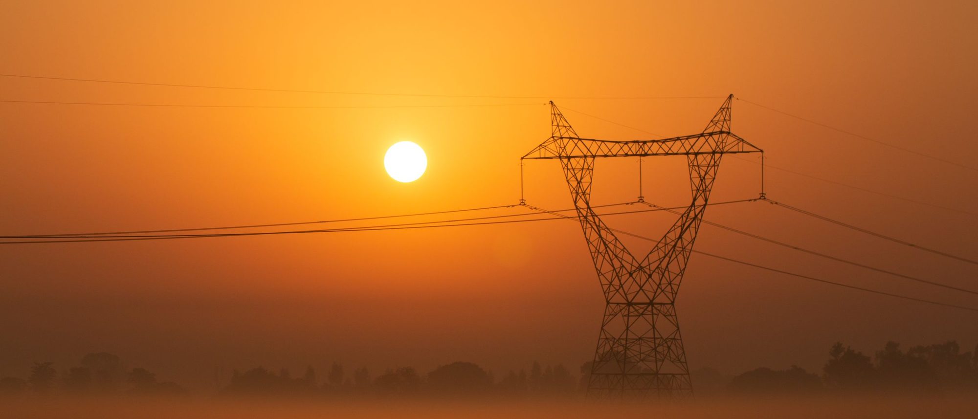 A power line tower in fog with the sun low in an orange sky.