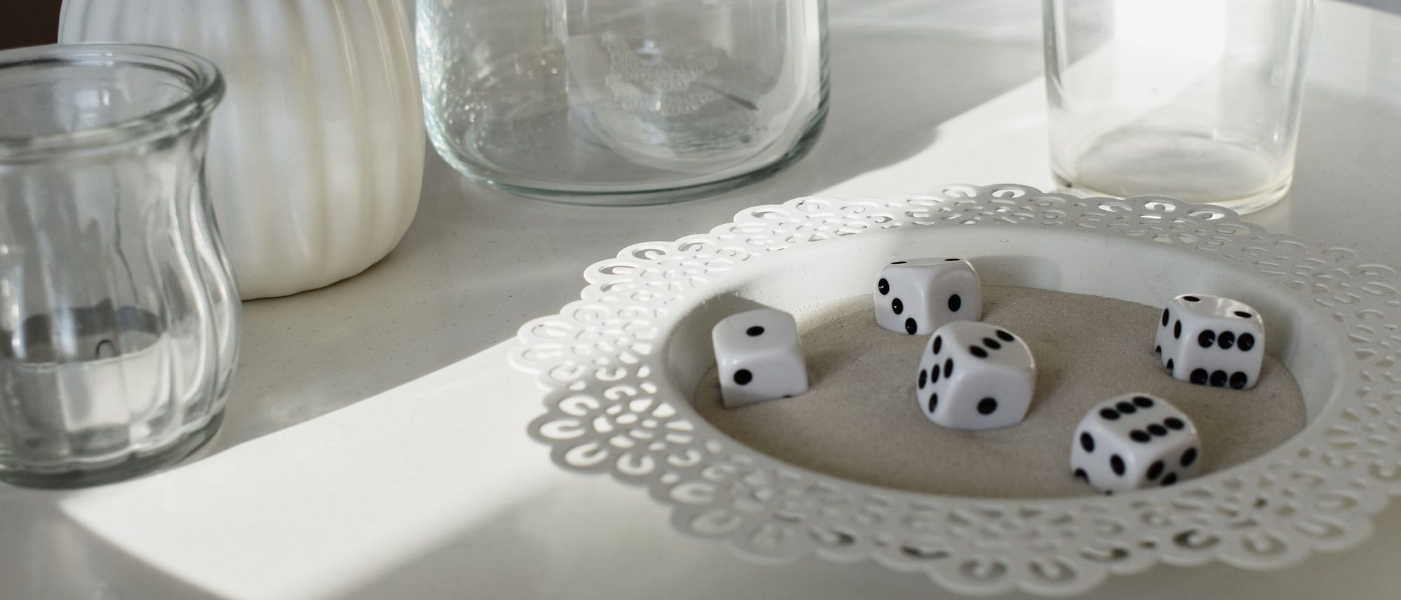 White and black dice sit among glassware and long shadows on a white table.