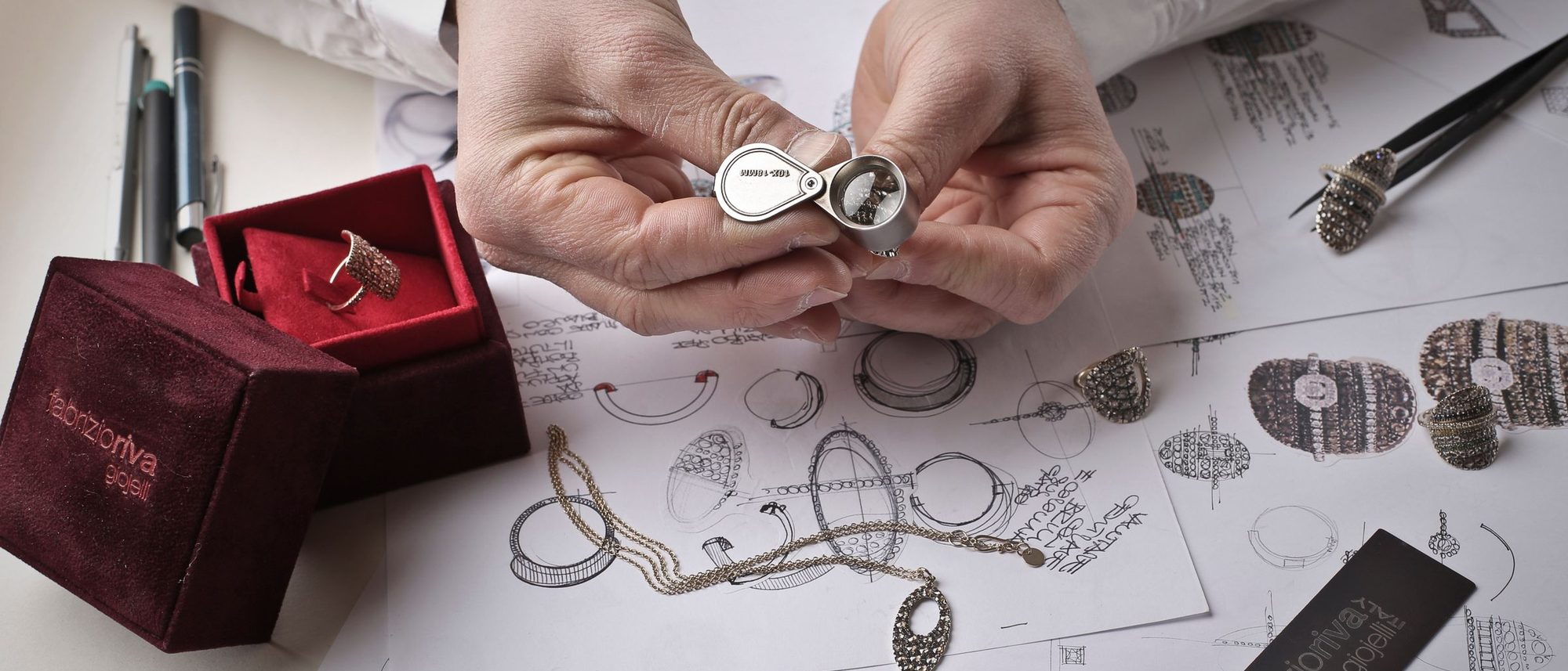 Two hands hold a ring under a jeweller’s loupe above a workbench scattered with sketches.