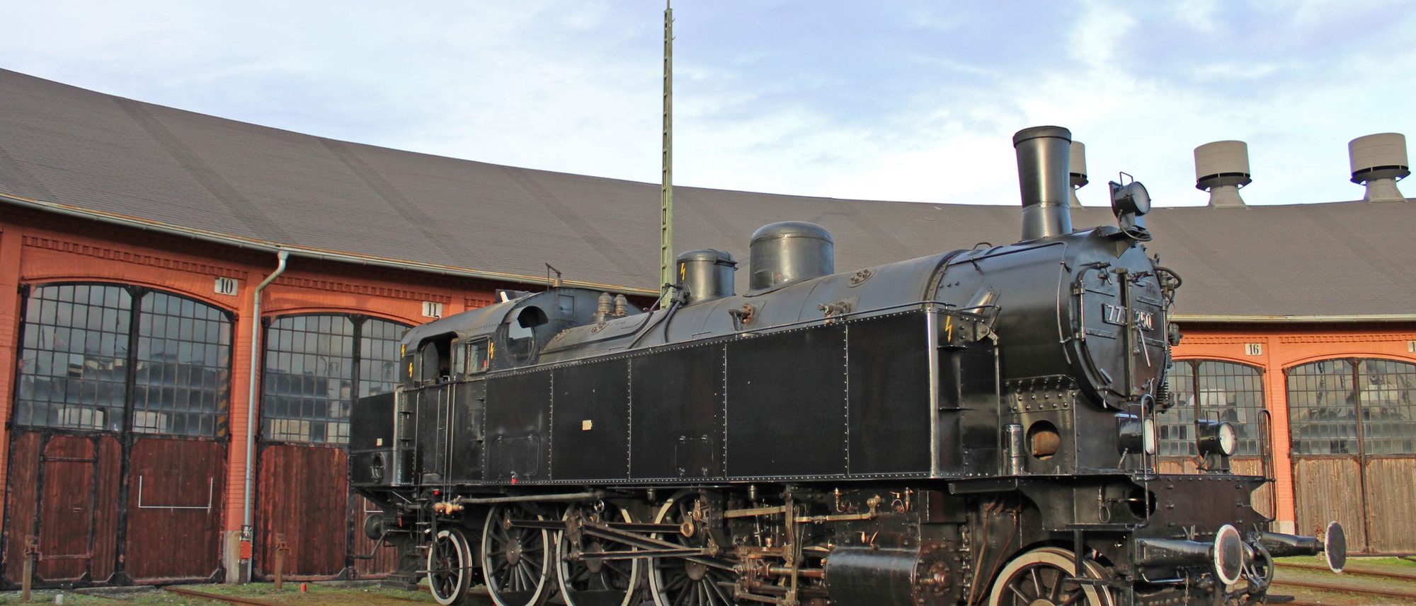 A preserved steam locomotive standing in front of a historic roundhouse.