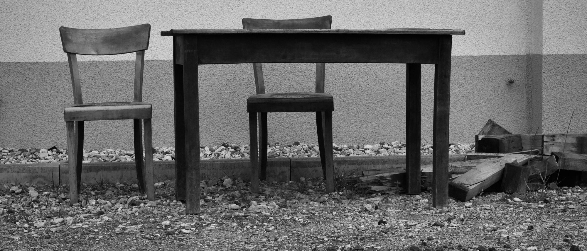Two worn wooden chairs and a small table stand against a wall outdoors in black and white.